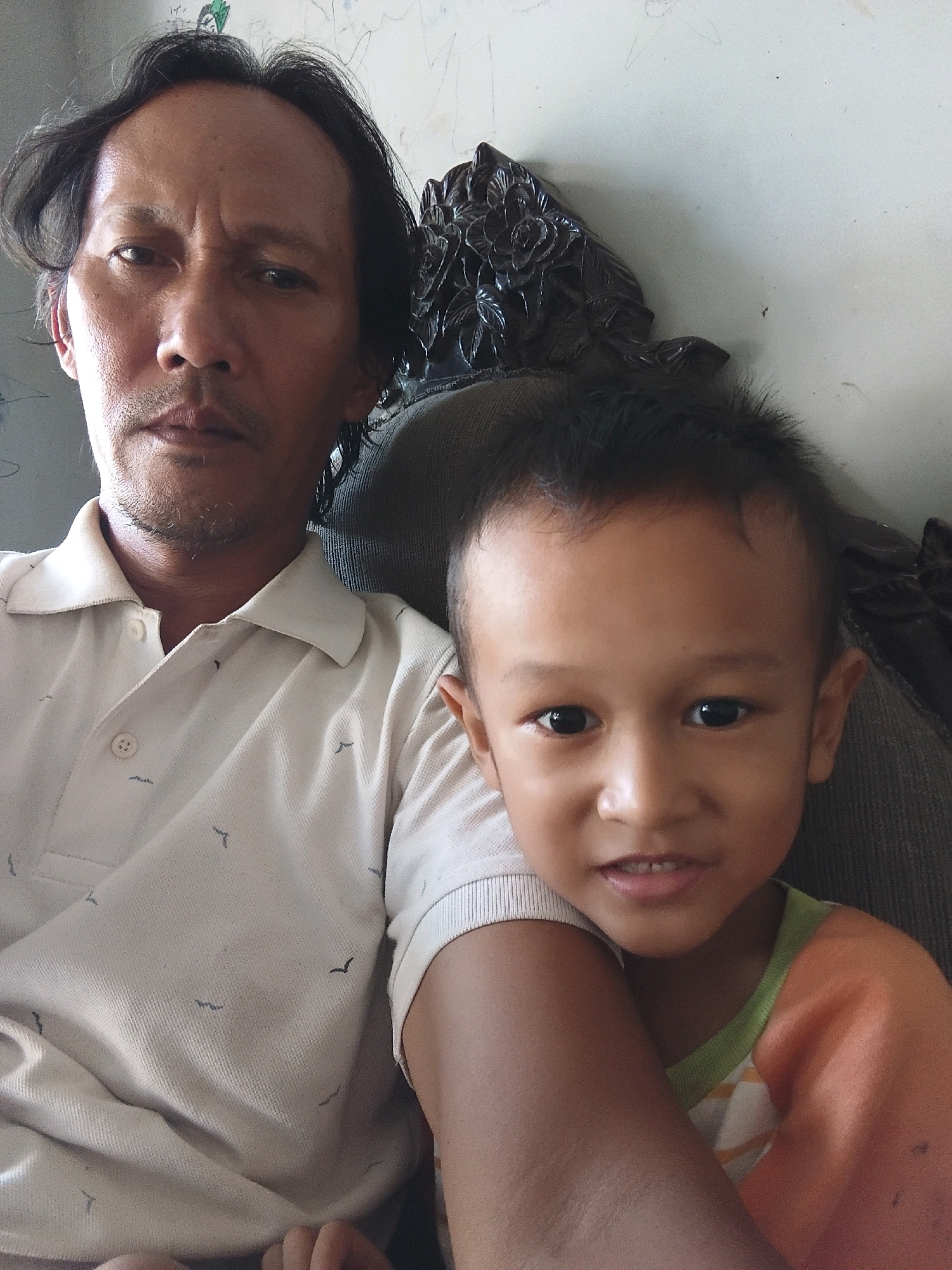 A father and son are photographed sitting together, likely on a couch. The father, on the left, is an adult male with dark hair, styled loosely, and a mustache and light stubble. He is wearing a light-colored polo shirt with a subtle bird pattern. His expression is somber, with his brow furrowed. The son, on the right, is a young child with dark hair and dark eyes. He is wearing an orange shirt with green trim. The child is looking directly at the camera with a slight smile. The background appears to be a wall with some faint scribbles or drawings. A dark, ornate object, possibly part of a chair or furniture, is visible behind the subjects. The lighting suggests an indoor setting, possibly a home in Cikal, Indonesia.