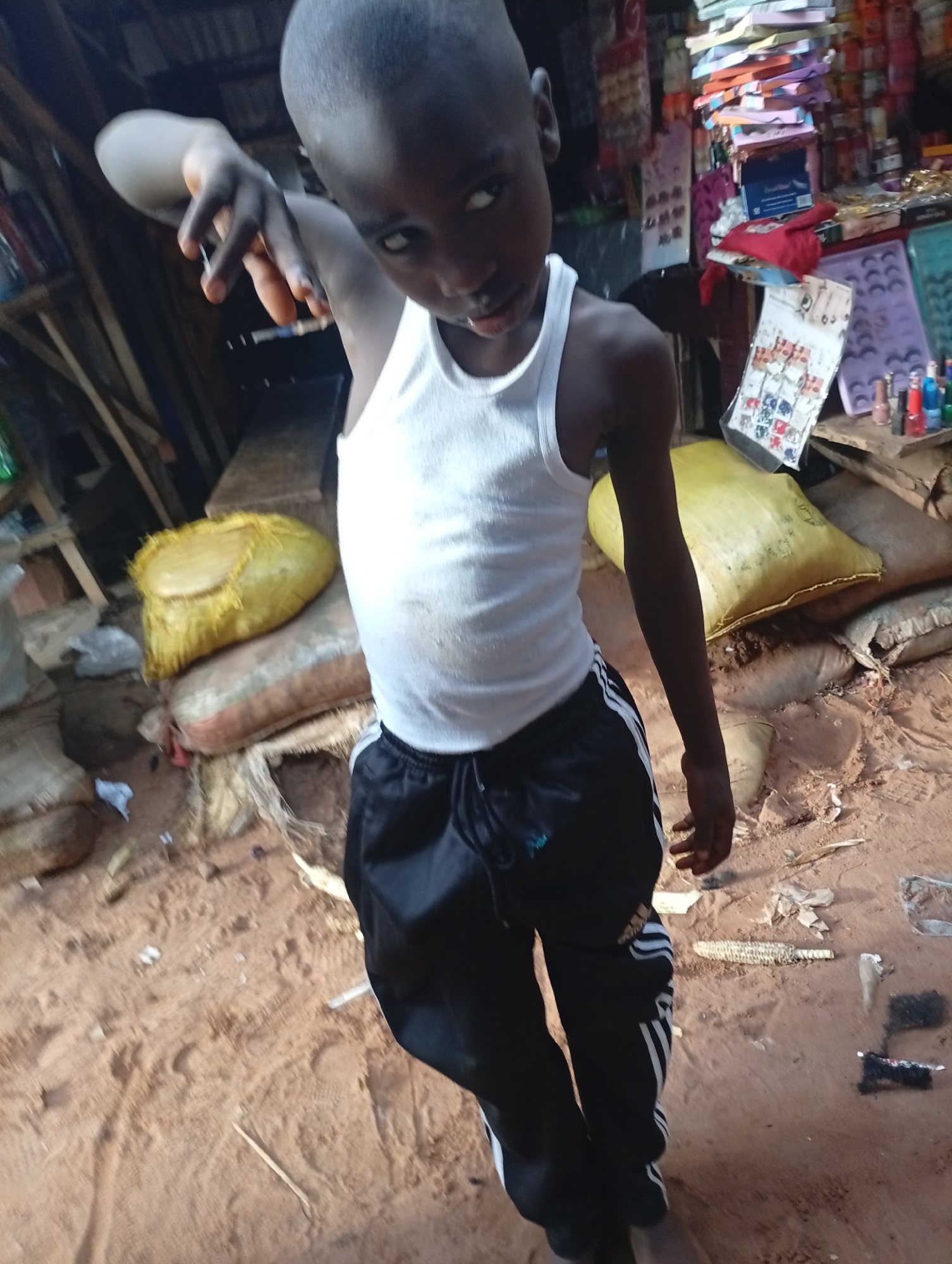 A young boy, likely pre-teen, stands in what appears to be a market or storage area in Nkpor, Nigeria. He is wearing a white tank top and black athletic pants with white stripes. His left arm is extended, and his hand is held up in a gesture, possibly for the camera. His expression is direct and engaging, with a slight smirk.
The background reveals a cluttered environment. Shelves filled with various goods, including what look like packaged items, toys, and cosmetics, are visible. In the foreground and mid-ground, there are several large sacks, some filled and others appearing to be empty or deflated, made of rough material. One sack is a bright yellow and covered in frayed fibers, resting on top of another sack with red and beige stripes. The ground is covered in reddish-brown dirt or sand, with some debris scattered around, including what looks like a cob of corn. The lighting suggests it is daytime, but the overall atmosphere is somewhat dim, perhaps due to the covered nature of the location or an overcast sky. The scene evokes a sense of everyday commerce and the environment of a local market in Nigeria. There are no discernible emotions beyond the boy's direct gaze.
