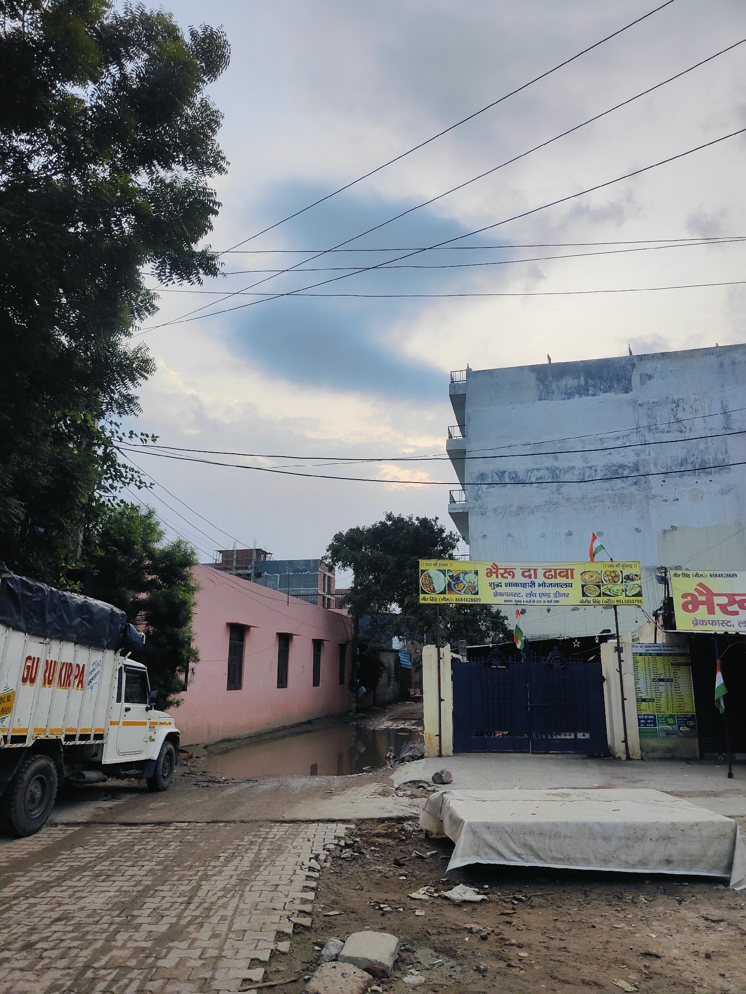 The image shows a street scene in Gurgaon, India, under a cloudy sky. In the foreground, a white truck with "GURU KIRPA" written on its side is parked. The road surface is a mix of cobblestones and dirt, with a large puddle of muddy water reflecting the surroundings. To the right of the truck is a pink building with dark windows. Further down the street, a large, light-colored building stands, adorned with Indian flags. A banner on this building reads "भैरू दा ढाबा" (Bhairu Da Dhaba) and lists menu items such as "ब्रेकफास्ट, लंच और डिनर" (Breakfast, Lunch, and Dinner). A dark blue gate is visible in front of this building. A white mattress covered with a sheet lies on the ground in the foreground. Trees with dark green foliage are present on the left and in the background. Power lines crisscross the sky. The overall impression is one of a somewhat unkempt urban environment.