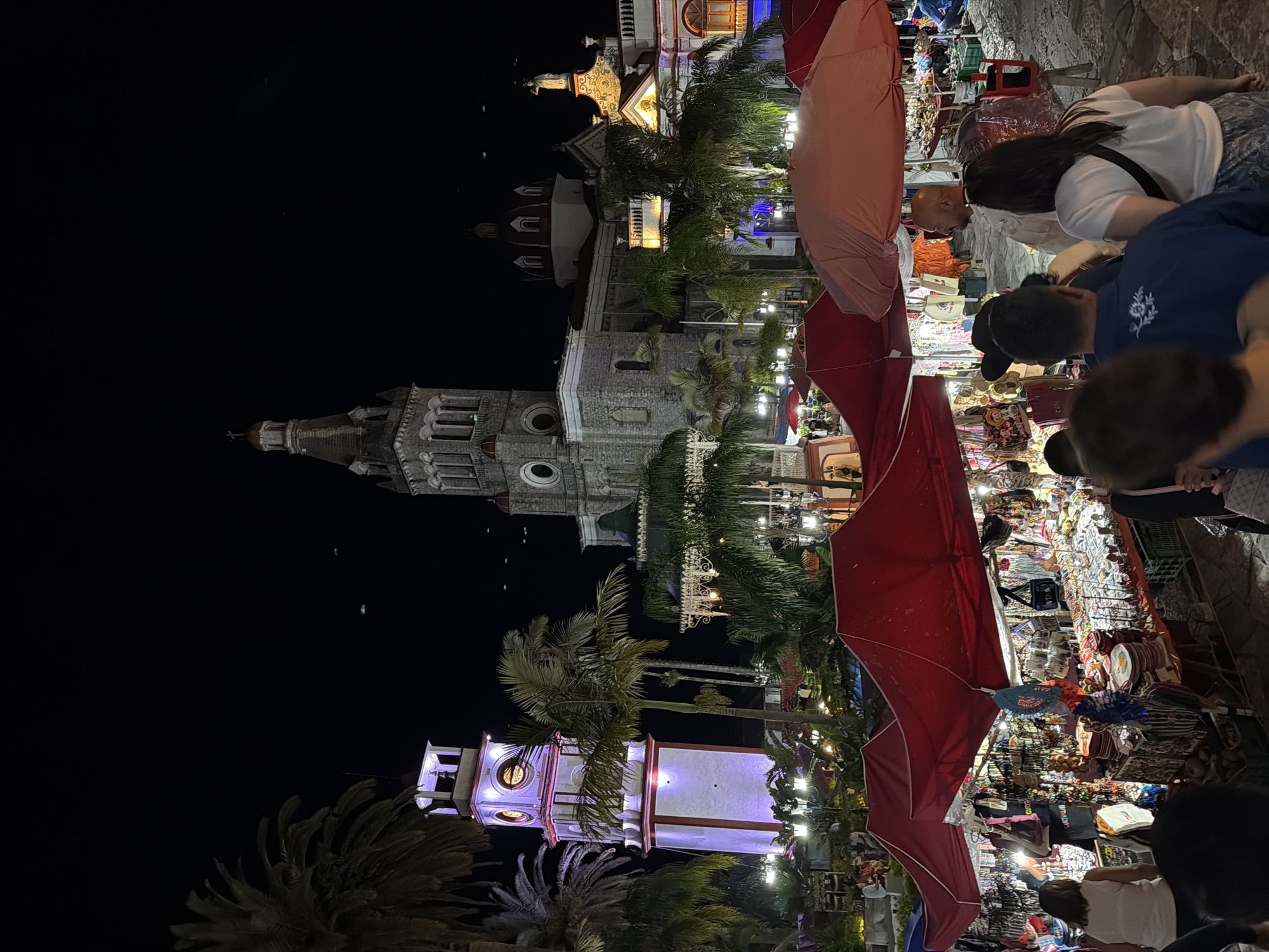 At night, the town square of Cuetzalan, Mexico, is alive with activity. A mix of locals and tourists browse the marketplace under red and pink awnings, examining souvenirs and handicrafts. A tall, illuminated clock tower and the nearby church, also lit up, punctuate the skyline against the night sky. People are looking at the goods being sold. Palm trees add a touch of tropical ambiance. The scene is a blend of commerce, architecture, and human interaction under the glow of artificial lights.
