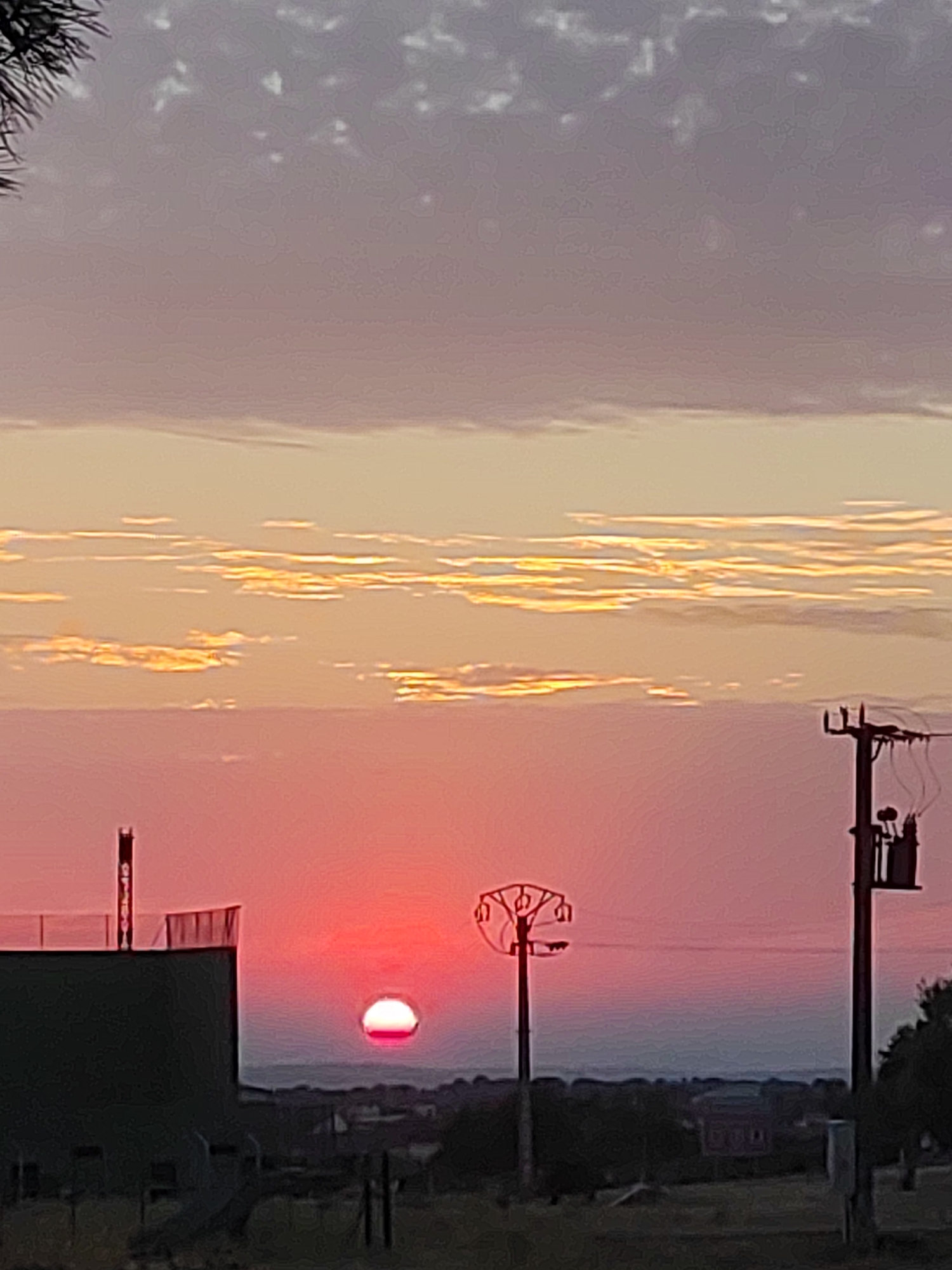 This is a vertical, medium-shot, outdoor photograph taken during sunset in the city of Sobradillo, Spain. The sky is filled with a gradient of colors from deep red at the horizon to light pink and then pale yellow with streaks of white clouds. A full, vibrant red sun is visible just above the horizon, partially obscured by the landscape. In the foreground, to the left, is a dark, curved structure with a tall, dark, vertical pipe extending from its top. To the right, a utility pole with a transformer box is visible. The middle ground shows a distant, hazy landscape with trees and some buildings. The overall scene depicts a tranquil sunset over the Spanish countryside.