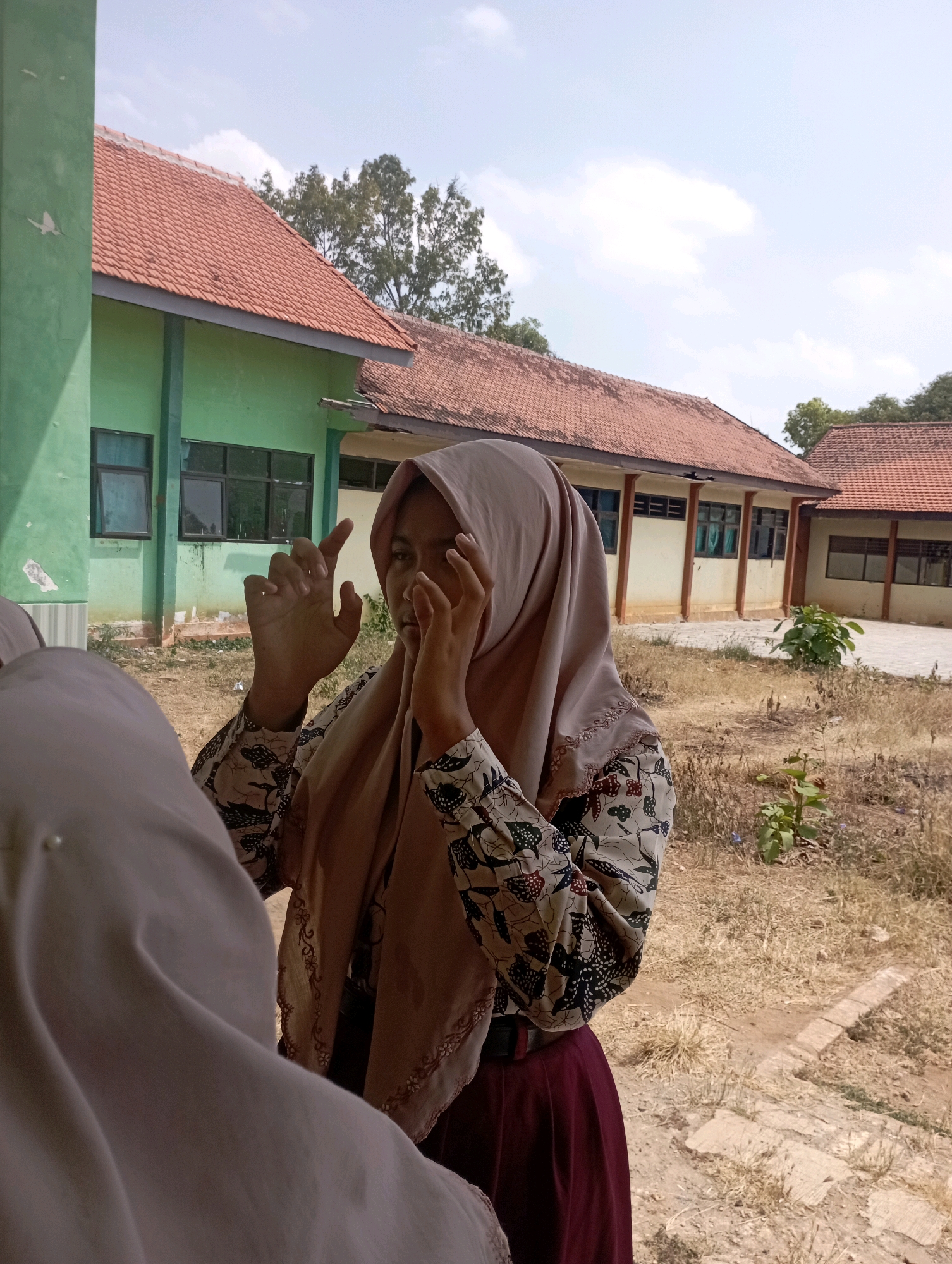 In Banyuurip, Indonesia, a young woman wearing a light brown hijab and a patterned blouse with dark red accents stands in front of a weathered green building with a red tile roof. She is partially obscured by another person, also wearing a light-colored hijab, whose shoulder and arm are visible in the foreground. The woman in the foreground is holding her hands up, with her fingers spread, near her face. The environment appears dry and arid, with patchy brown grass and dry vegetation surrounding a paved area. The building's architecture suggests it might be a school or similar public facility. The sky is blue with scattered white clouds.
