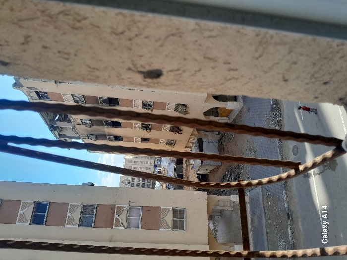 A low-angle view through a weathered metal railing shows damaged buildings across a street. The buildings are multi-story with decorative window surrounds. A figure in red is walking along the street. Debris litters the area. The sky is blue, visible between buildings. A "Galaxy A14" watermark is visible on the right side of the frame. The scene depicts urban decay.