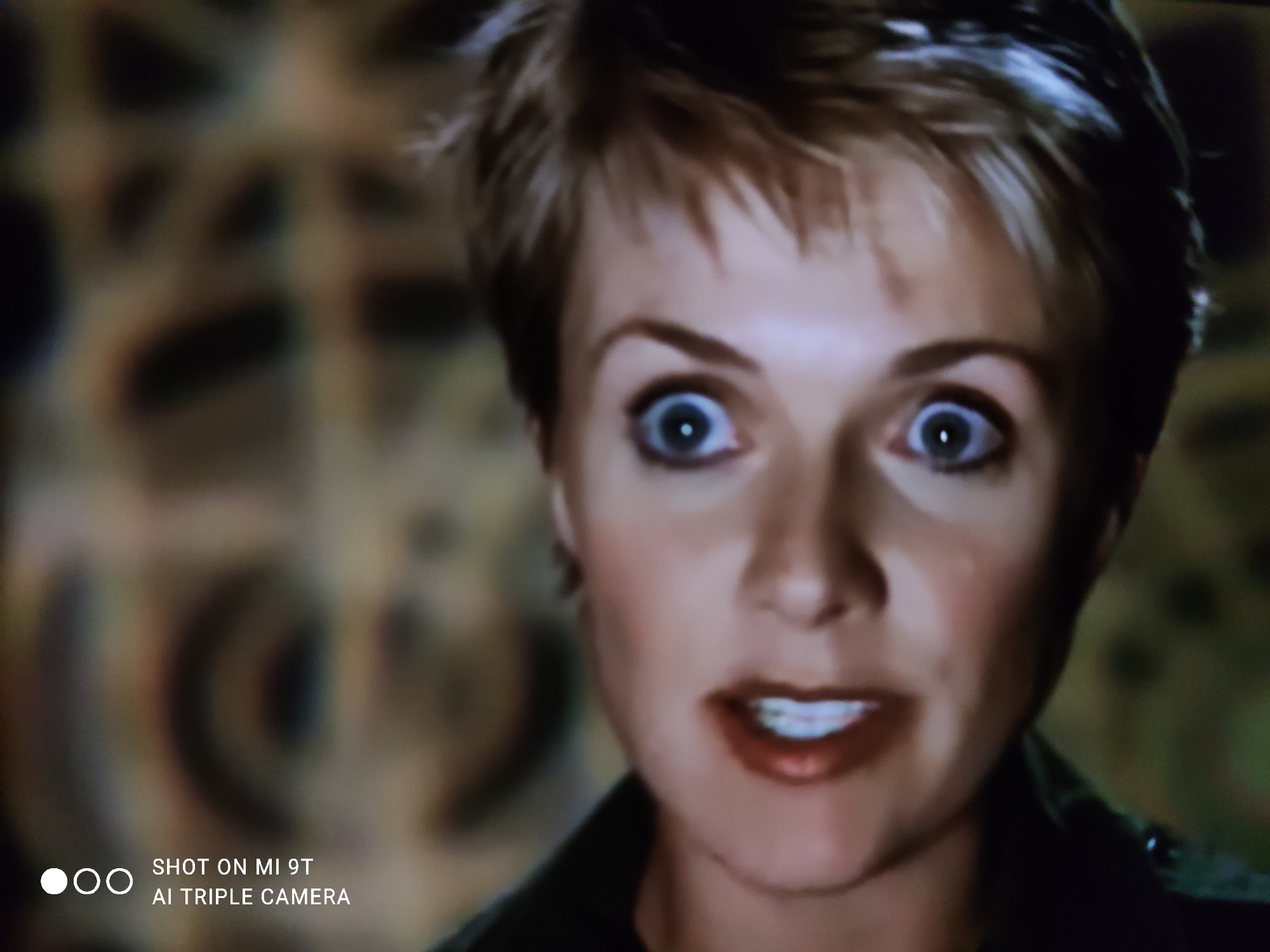 The image is a close-up, slightly blurred, photograph of a woman's face. She has short, spiky blonde hair and her eyes are wide open, looking directly at the camera with a surprised or shocked expression. Her mouth is slightly open, revealing her teeth, and she is wearing dark lipstick. The background is out of focus and appears to be a patterned fabric, possibly a tapestry or wallpaper, with shades of brown and beige. There is a watermark in the lower left corner that reads "SHOT ON MI 9T AI TRIPLE CAMERA". The scene does not provide any discernible details related to the city of Khazar Raion, Azerbaijan.