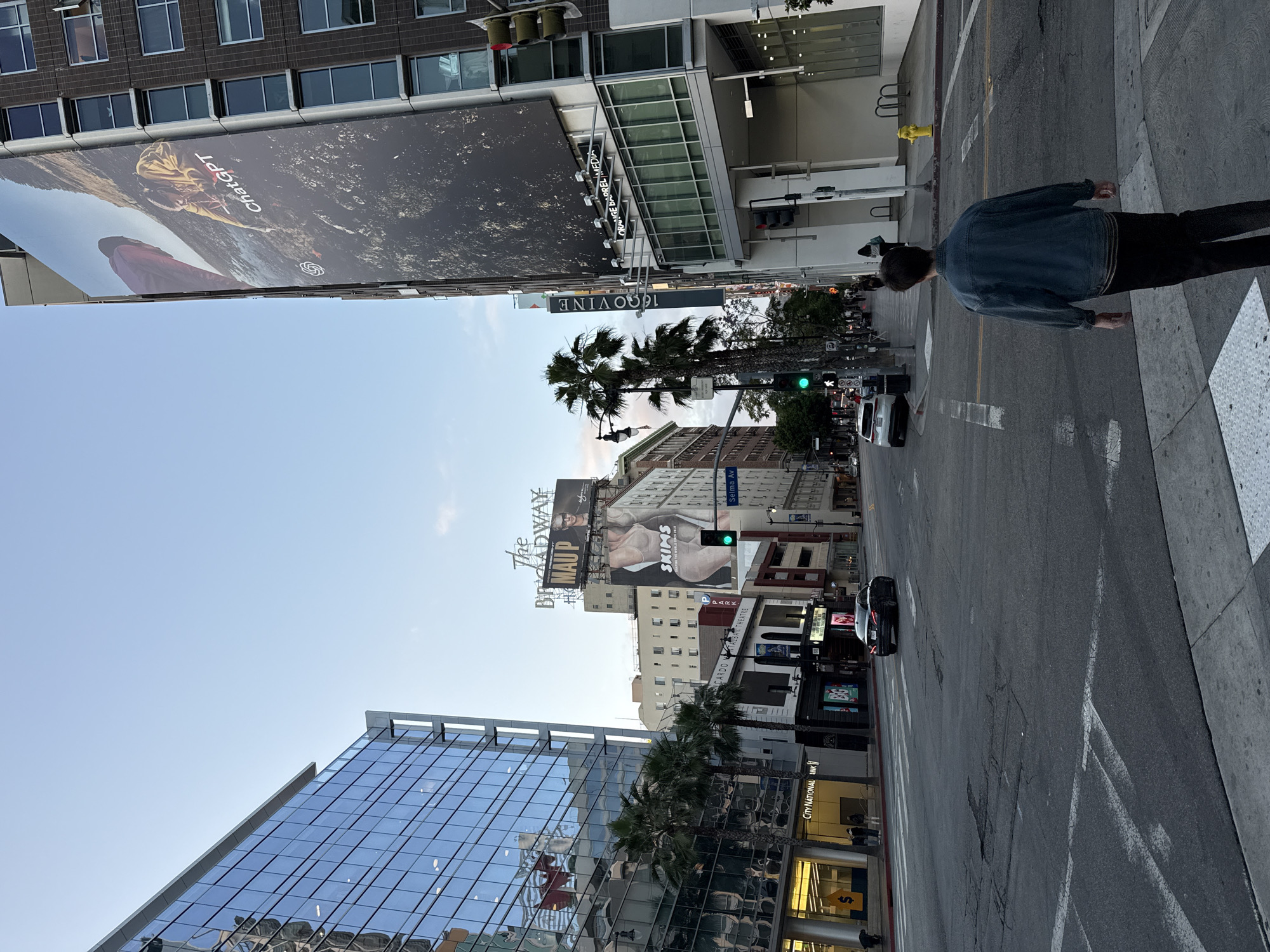 The image depicts a vibrant street scene in Los Angeles, captured during what appears to be the late afternoon or early evening. The sky is clear, with a soft, diffused light, and some artificial lights on buildings are beginning to glow.
In the foreground on the right, a person wearing a denim jacket and dark pants walks along a sidewalk, facing away from the camera. The street, marked with pedestrian crossings and traffic lanes, stretches into the distance, with a few cars visible, including a white vehicle stopped at a green traffic light. Palm trees are interspersed among the buildings, a signature feature of the city.
The street is flanked by various buildings, ranging from a modern glass-fronted structure on the left that reflects the sky, to older, multi-story buildings further down the street. Large billboards and signs are prominent. A massive billboard on a building on the left features the text "ChatGPT" with an accompanying image of people. Further down on the right, another large billboard advertises "SKIMS" with a model. Other visible text includes "The BROADWAY" and "MAUP" on a building sign, "1600 VINE" on a tall sign, "ORANGE BARREL MEDIA" on a building's signage, and "CITY NATIONAL BANK" on a storefront. A small street sign reads "Selma Av".