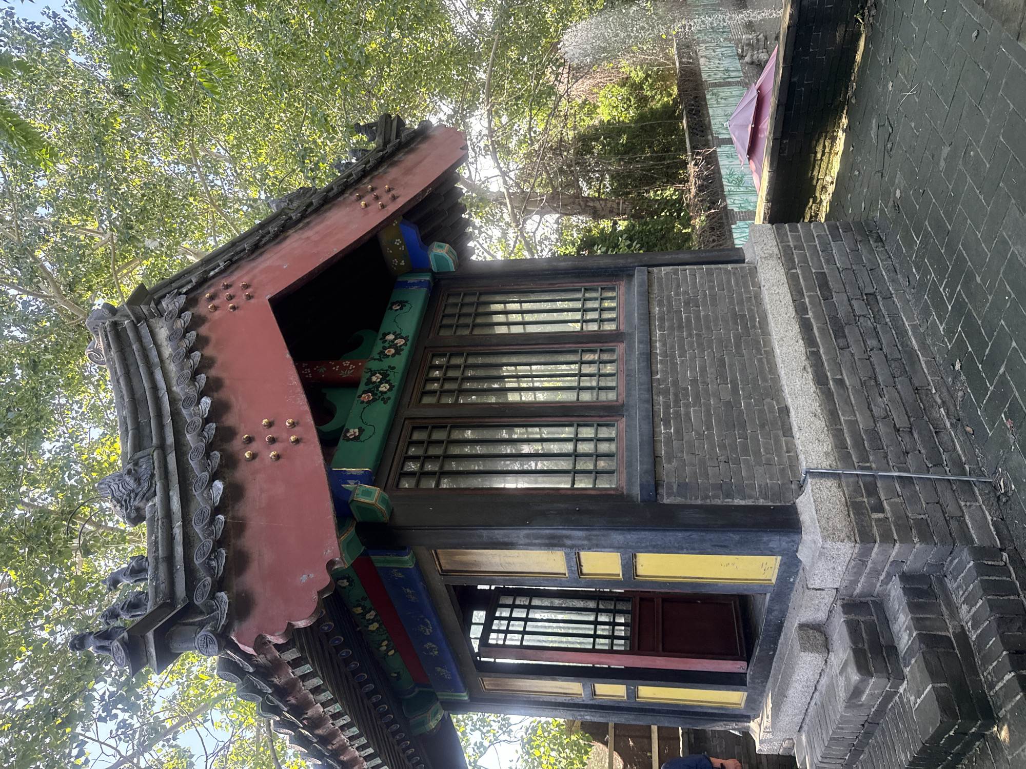 The image shows a traditional Chinese pavilion situated in an outdoor setting. The pavilion has a multi-tiered, dark-tiled roof with decorative eaves painted in red, green, and blue. The roof is adorned with small golden studs and ornate sculptures, including what appear to be small guardian lions and a dragon head. The walls of the pavilion are constructed with dark grey bricks, and it features large, multi-paned windows with dark frames. One side of the pavilion has an open doorway, revealing a red door and more windows. The entrance is accessed by a short set of brick steps. The pavilion is surrounded by paving stones and a low brick wall. In the background, lush green trees with dense foliage are visible, and sunlight filters through the leaves. To the right of the pavilion, a small, multi-tiered red umbrella-like structure is partially visible behind the wall. The scene appears to be located in a park or garden, likely within the city of Qinhuangdao, China. There are no people clearly depicted in the image, although a hint of a person wearing dark clothing is visible on the far left. The overall impression is one of serene, traditional architecture set against a natural backdrop.