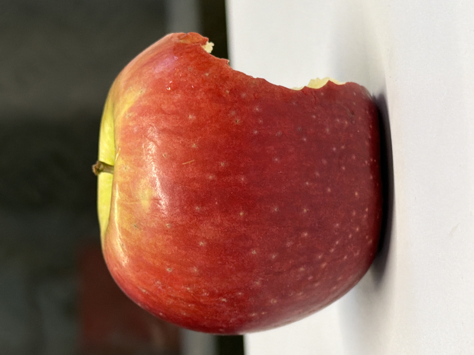 A red apple with a bite taken out of it is positioned on a white surface. The apple is mostly red, with some yellow and lighter red patches near the stem. The bite mark is on the right side of the apple, revealing the lighter flesh inside. The background is dark and out of focus, with a hint of a red object visible to the left. The scene appears to be indoors.