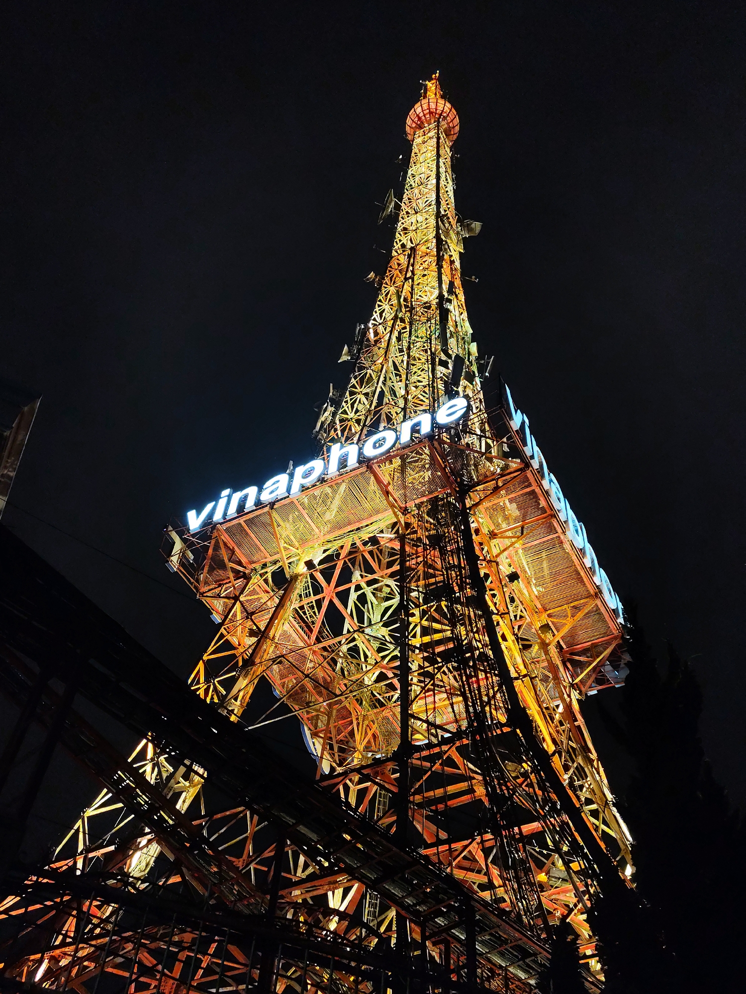 The image depicts the illuminated Vinaphone communications tower at night in Da Lat, Vietnam. The tower is primarily orange and yellow, constructed with a lattice-like metal framework, and has the word "vinaphone" illuminated in blue-white lettering on its upper levels. The tower rises vertically from the bottom of the frame towards the upper center. There are dark utility cables in the foreground that intersect the view of the lower portion of the tower. The background is a dark night sky, with faint hints of buildings and trees visible on the edges of the frame.