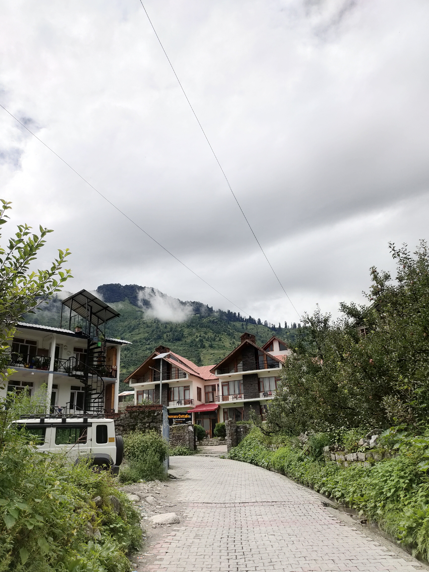 The image depicts a view of a road leading towards a couple of buildings with mountainous terrain and clouds in the background, possibly in Burwa, India. The road is paved with light-colored bricks and is lined with lush green vegetation on the right. On the left, there is a white SUV parked next to a white multi-story building featuring an external spiral staircase. Further back, another building with a reddish-tiled roof and stone accents is visible. The mountains in the background are covered in green trees, with some patches of white clouds obscuring the upper slopes. The sky is overcast with heavy, gray clouds, suggesting a cloudy or potentially rainy day. There are power lines stretching across the sky, contributing to the somewhat utilitarian aspect of the scene despite the natural beauty of the landscape. No people are visible in the image.