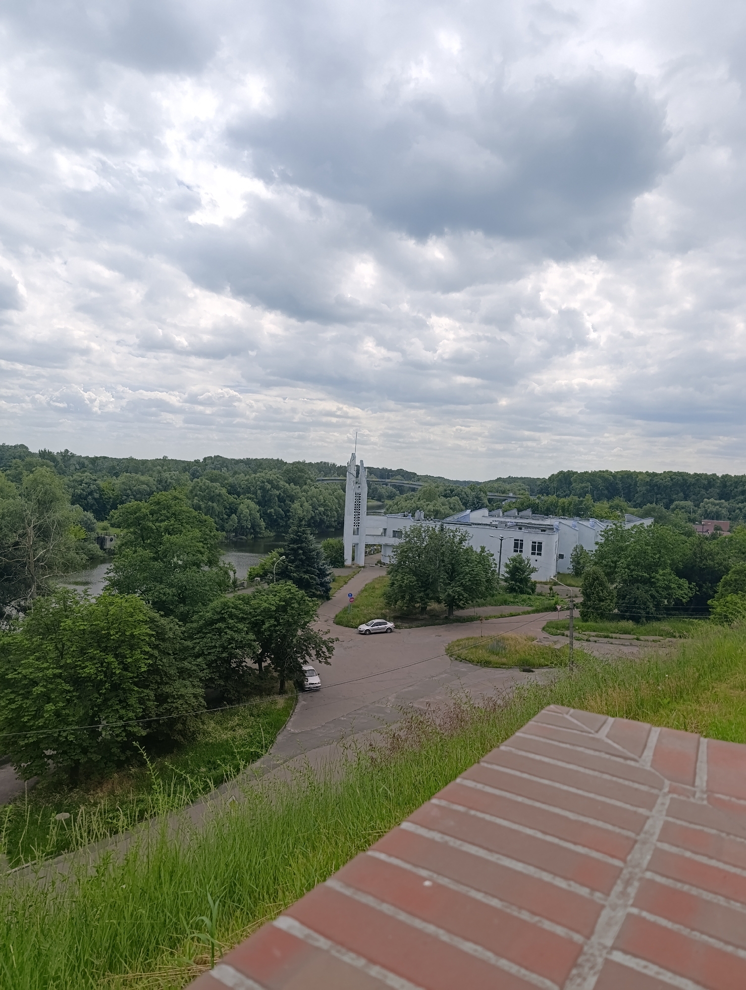 A high-angle, wide shot shows a cloudy sky above a cityscape with a body of water and lush green trees. In the foreground, a red brick wall partially obscures the view. A white, modern building with a tall, slender tower stands to the right of a body of water. Further back, a long, white building with a sloped roof is visible, surrounded by trees. Several cars are parked or driving on paved roads and pathways. The environment appears to be a park or urban area with significant natural vegetation. This scene is located in Chernihiv, Ukraine, specifically it is the territory of the Chernihiv National Technological University on the banks of the Stryzhen River.