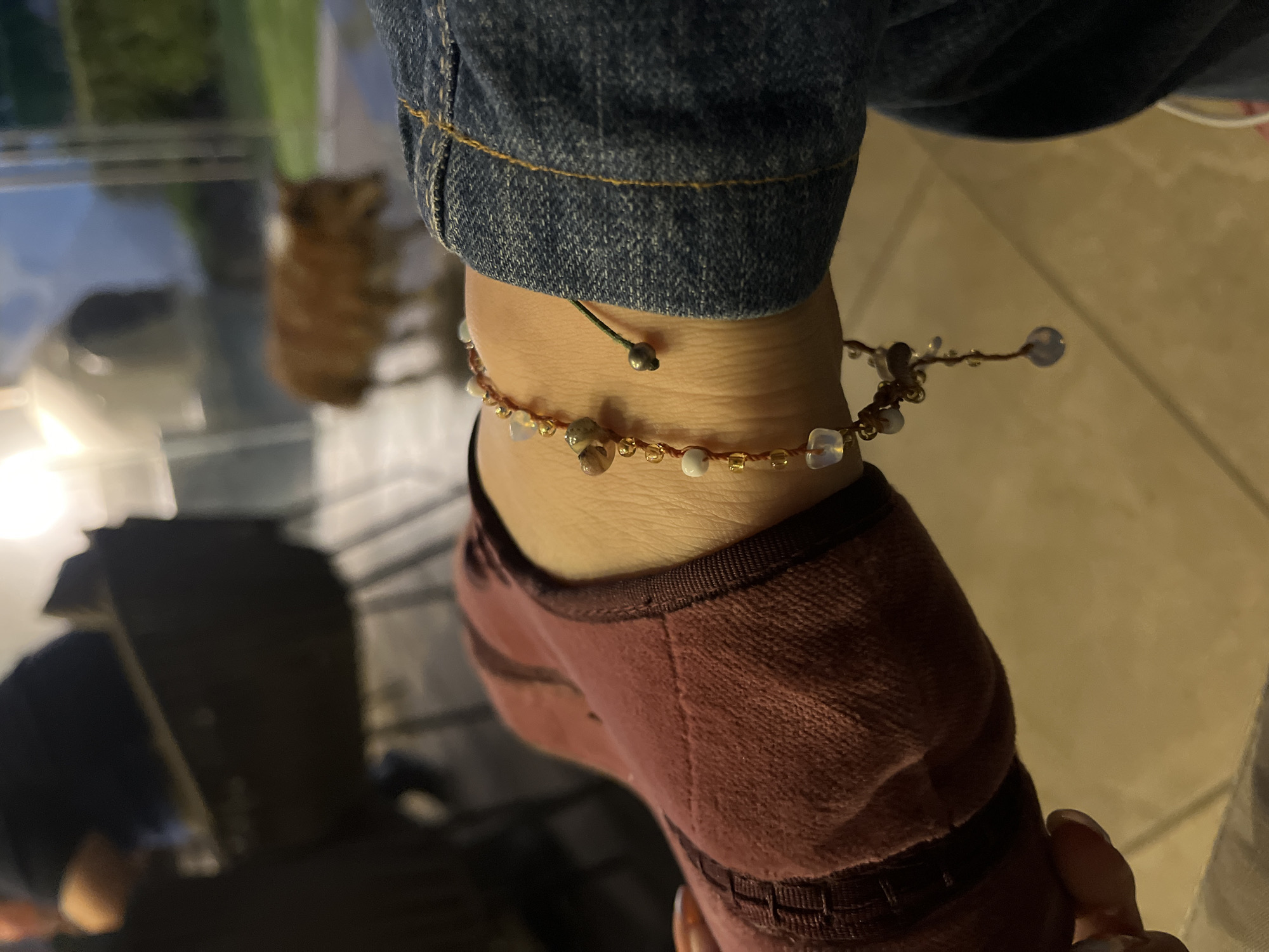 A close-up shot shows a person's ankle adorned with a beaded anklet. The anklet consists of small, light-colored beads and a brown cord. The person is wearing a reddish-brown sock and denim jeans. The setting appears to be indoors with a tiled floor. The background is blurred, but hints of furniture are visible. The location in the city of San Bernardino Tlaxcalancingo, Mexico, is not determinable from the image.