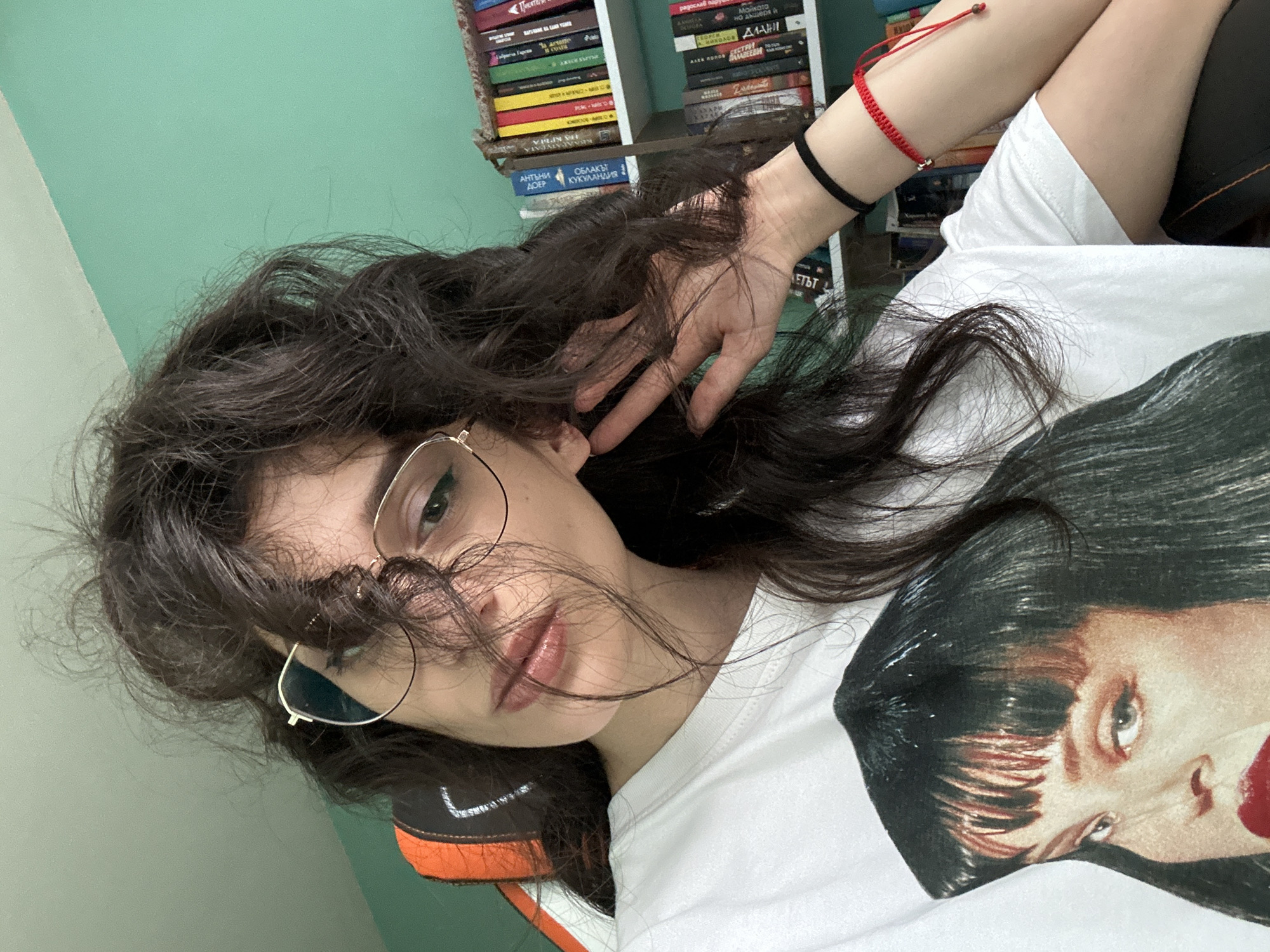 The media file shows a close-up, rotated selfie of a young person with long, dark, wavy hair and glasses. The person is wearing a white t-shirt with a graphic of a woman's face printed on it. The background features a mint green wall and a bookshelf filled with books. On the person's left wrist, there are two bracelets: a black one and a red string bracelet. The person's right hand is partially visible, resting on their hair. The lighting suggests it is daytime. The scene appears to be indoors, possibly a bedroom or study, given the bookshelf. The person's expression is somewhat casual, with their gaze directed towards the camera. There is no discernible weather or time of day information beyond the general indoor lighting. There is visible text on the spines of the books on the shelf, but it is not legible. The location context provided is Sofia, Bulgaria.