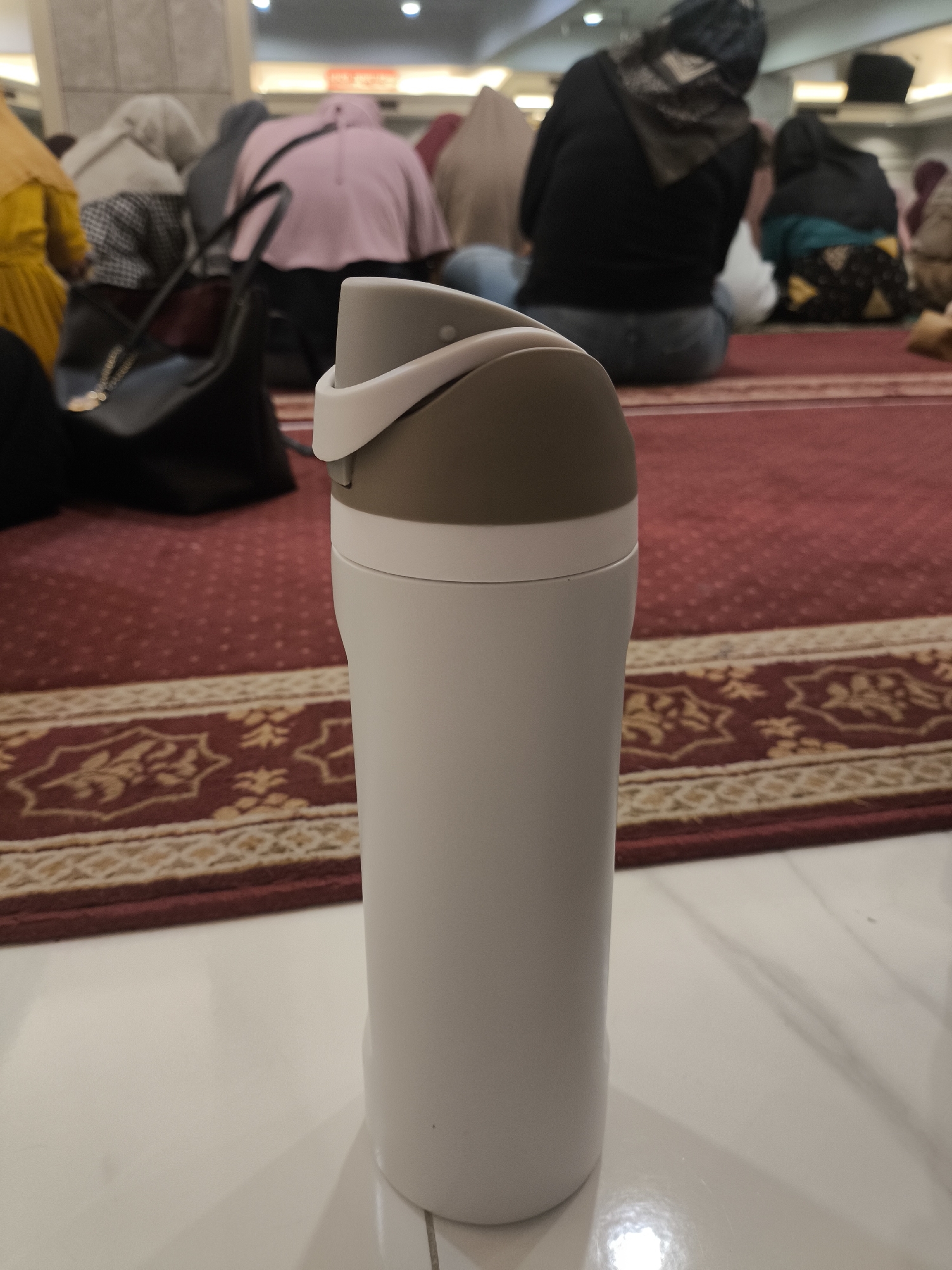 In the city of Grogol, Indonesia, a white and brown tumbler stands on a white tiled floor in the foreground. Behind the tumbler, a red carpet with a patterned border stretches across the ground. Several individuals, their backs turned to the camera, are seated on the carpet, facing away from the viewer. They appear to be in a place of worship, possibly a mosque, as some are wearing headscarves and the setting suggests a communal gathering. The lighting in the background indicates an indoor environment.