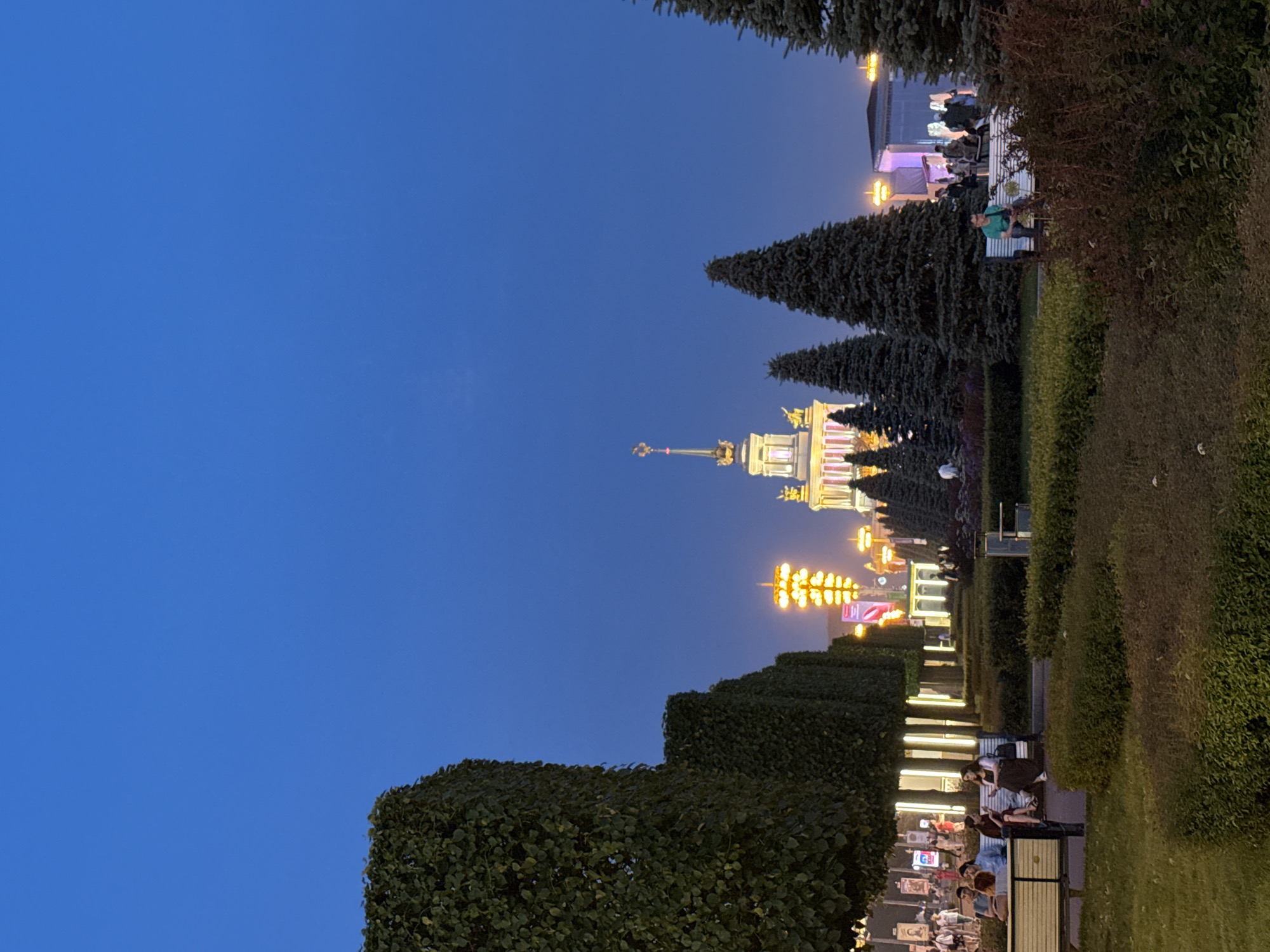 The image depicts a twilight scene at the All-Russian Exhibition Center (VDNKh) in Moscow, Russia. A tall, ornate building with a golden spire is visible in the background, illuminated by lights, along with other illuminated structures and tall, slender lights lining a pathway. In the foreground, a manicured park area features rows of well-maintained hedges and trees, including several tall, dark green fir trees. People are seated on benches and walking along pathways, suggesting a leisurely evening atmosphere. The sky is a deep, clear blue, indicating the time of day is just after sunset.