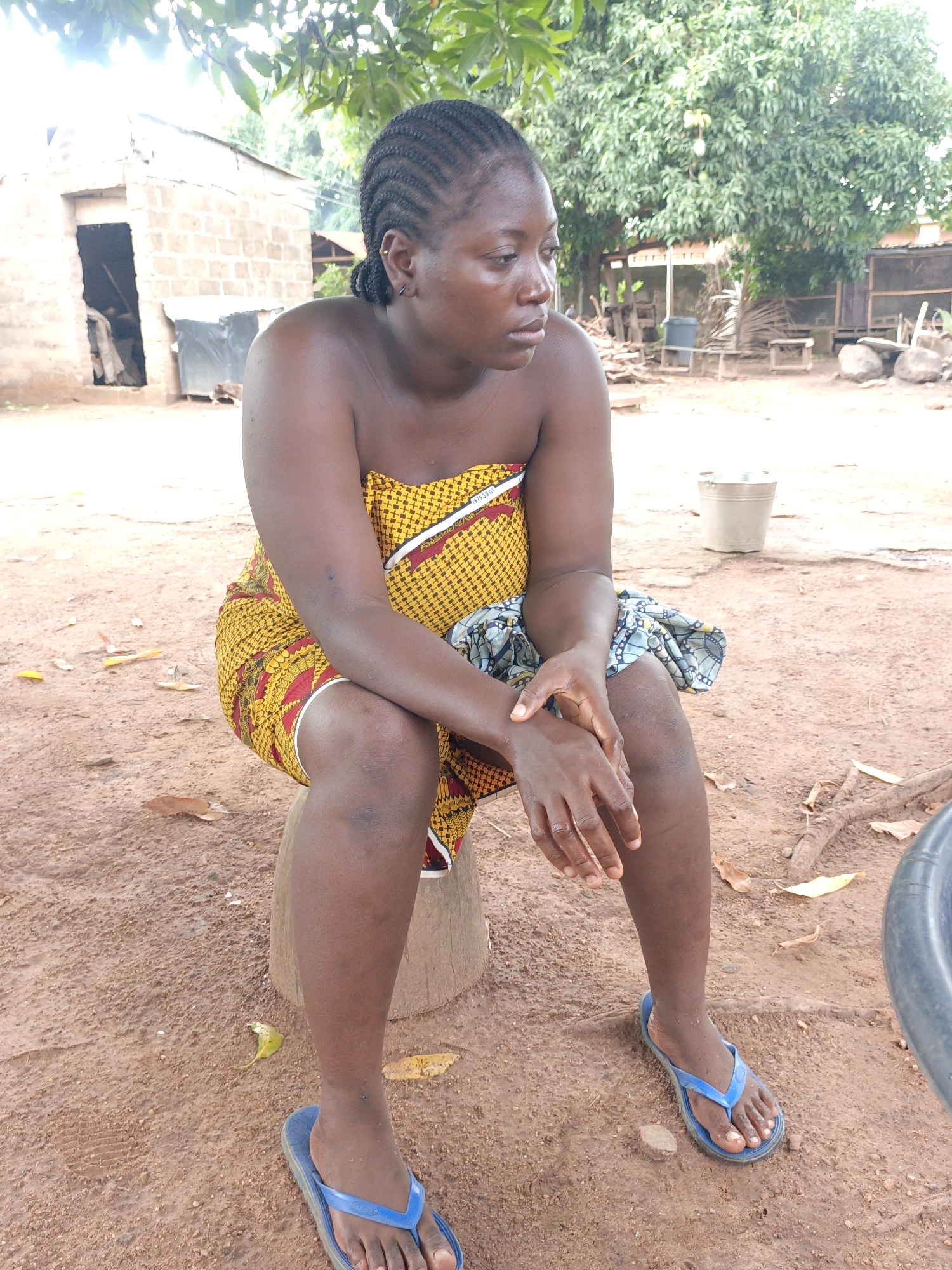 A woman is sitting on a stool in an outdoor setting. She is wearing a yellow and red patterned cloth wrapped around her body and blue flip-flops. Her hair is styled in braids. She has a serious or pensive expression and is looking to her right. Her hands are clasped together in her lap.
The background includes a brick structure, possibly a shed or small building, along with trees and other vegetation. There is a bucket and some wooden structures visible in the distance. The ground appears to be dirt or clay. The location is a rural environment.
