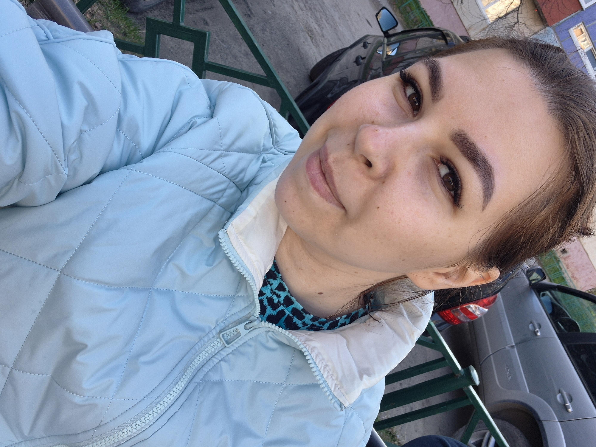 A woman is taking a selfie outdoors. She has brown hair pulled back and is wearing a light blue quilted jacket over a teal and black top. A section of green metal railing is visible in the foreground. Behind the woman and railing, two parked cars are partially visible. A portion of a building, painted with blue and red panels, is also in the background. The location is likely the city of Tobolsk, Russia.