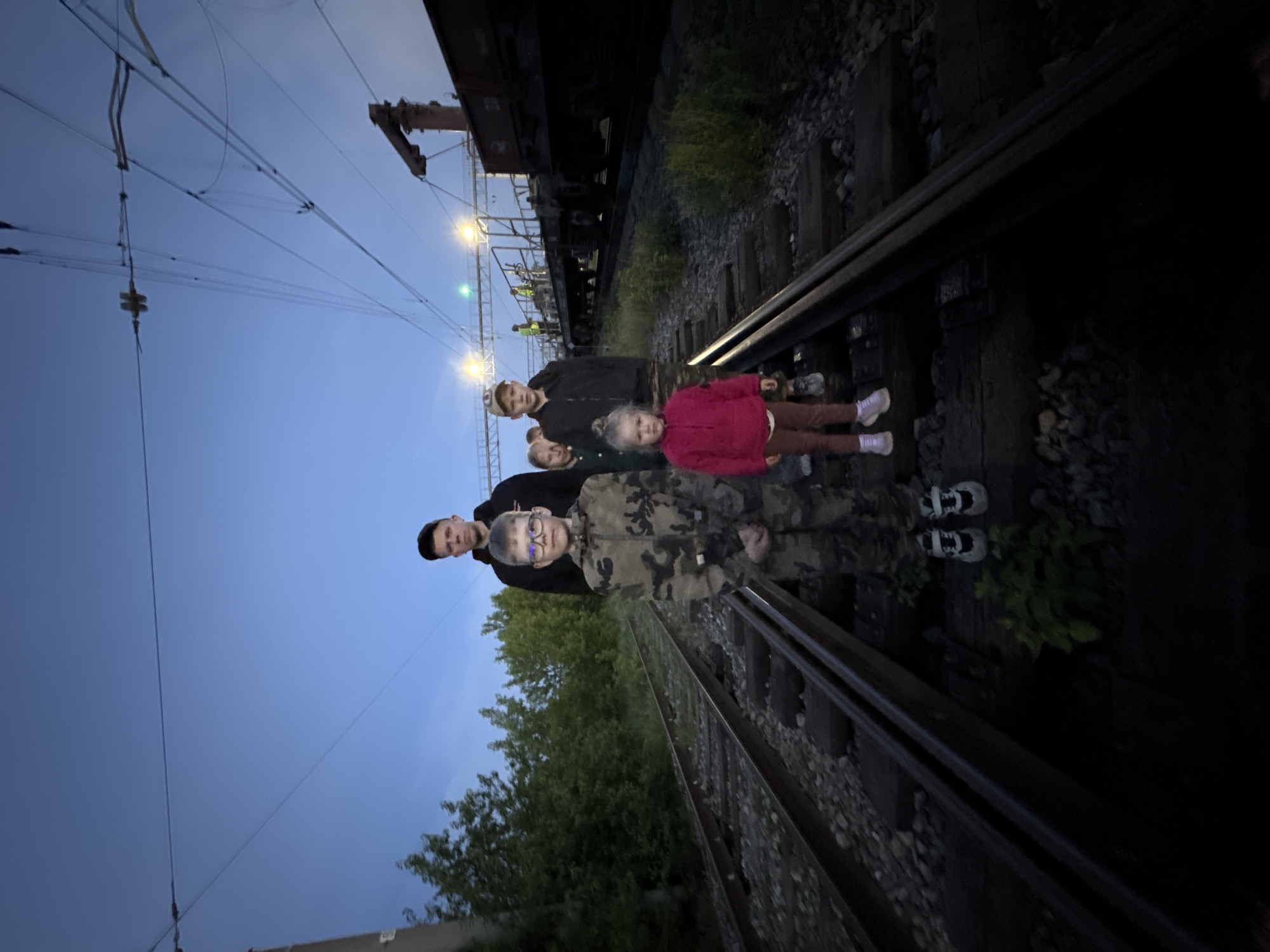 The image shows four people standing on railroad tracks in Ферма, Russia. From left to right, the people appear to be: a male in a black shirt, a male in a black jacket and a baseball cap, a young female in a pink jacket, and a young male in a camouflage jacket wearing eyeglasses. A freight train is parked in the background, along with overhead electrical lines. The area near the tracks has scattered gravel and some vegetation. The sky is a pale blue.
