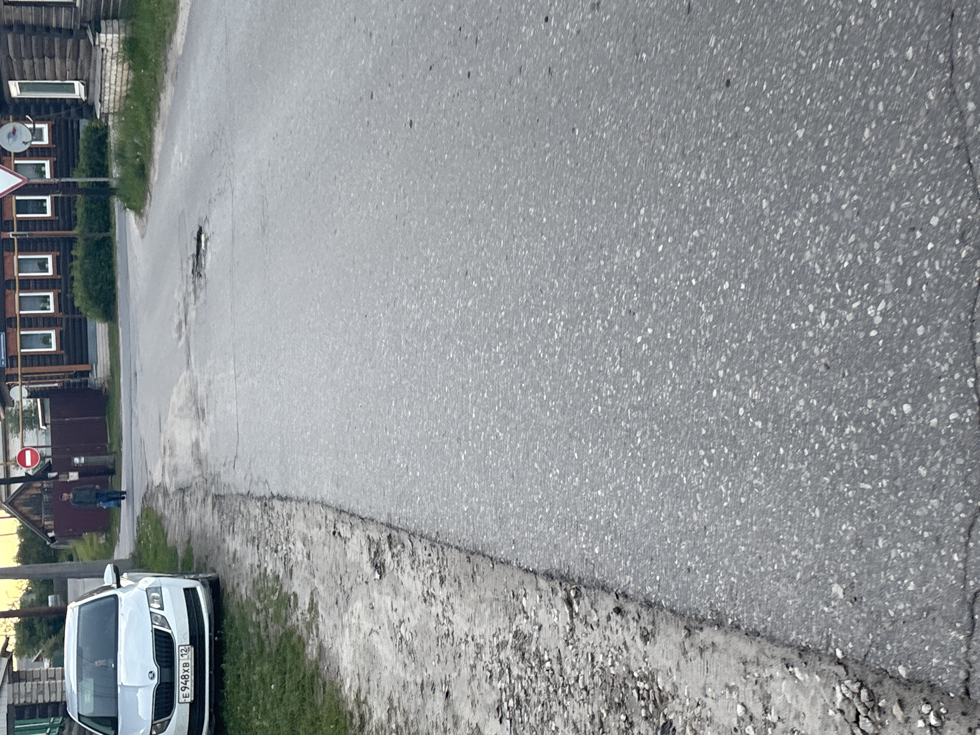 The image displays a street scene, likely in a rural or semi-urban setting, oriented horizontally due to the image's rotation. An asphalt road dominates the center and right side of the frame, showing signs of wear with visible cracks and a prominent pothole further down. The road is bordered by an unpaved shoulder composed of dirt and gravel.
On the left side, a white Skoda car is parked on the grassy edge next to the road. The car's license plate, "E 948 XB 12", is clearly visible, indicating registration in the Mari El Republic, Russia. Behind the car and along the street are several buildings, predominantly constructed of wood with multiple windows. One building has a satellite dish mounted on its side. Further down the street, a red circular road sign with a white horizontal bar, typically indicating "No Entry" or "Do Not Enter," is visible. A single person is standing in the distance near the buildings.
The scene suggests a calm, ordinary moment. The lighting appears soft and diffused, characteristic of an overcast day or early morning/late afternoon. No specific activity is actively taking place; it's a static view of a residential or village street. The architecture of the buildings and the style of the road sign align with typical visuals found in Russia.