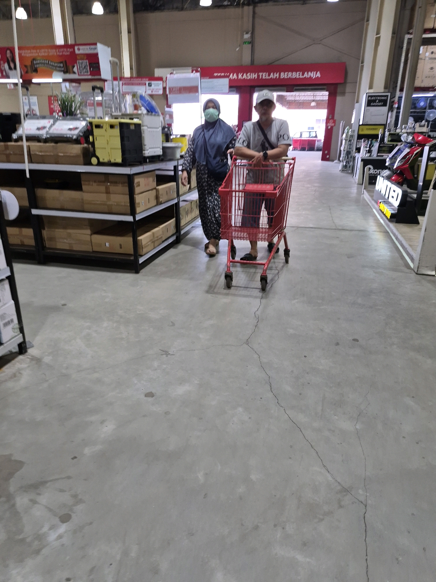 In an indoor commercial setting, a man and a woman are walking side-by-side, pushing a red shopping cart. The man, wearing a white cap and a grey t-shirt, is positioned on the right, while the woman, wearing a blue hijab and a patterned dress, is on the left. They appear to be customers in a retail store. The environment features concrete flooring, tall shelving units stocked with boxes and merchandise, and overhead lighting. A sign in the background reads "TERIMA KASIH TELAH BERBELANJA", which translates to "THANK YOU FOR SHOPPING". This scene is consistent with a retail environment such as a Lotte Mart hypermarket, commonly found in areas like East Jakarta, Indonesia, known for their wide aisles, extensive product displays, and customer service signage.