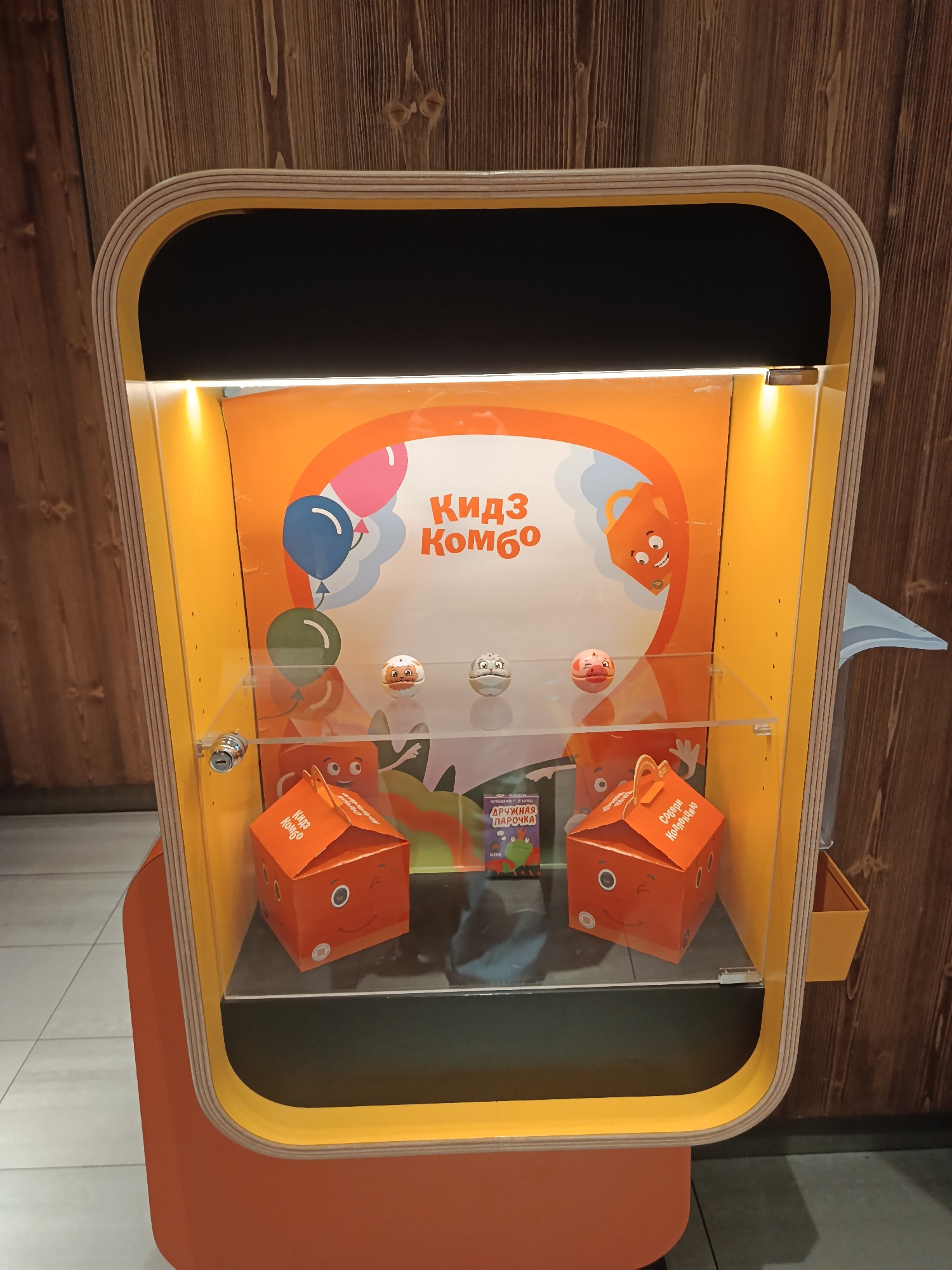 The image shows a "Kidz Combo" display case, likely in a fast-food restaurant in Moscow, Russia. The case has a rounded rectangular frame, constructed from layered plywood with a yellow inner and orange outer surface. The interior is brightly lit, with an orange background featuring cartoon characters, balloons, and the words "Кидз Комбо" in Cyrillic lettering. A clear acrylic shelf contains three small, round toys and two orange, cube-shaped "Kidz Combo" boxes. A children's book with Cyrillic text is positioned between the boxes. The case is likely part of a promotional display for children's meals. The background is a wood-paneled wall and a white tiled floor.