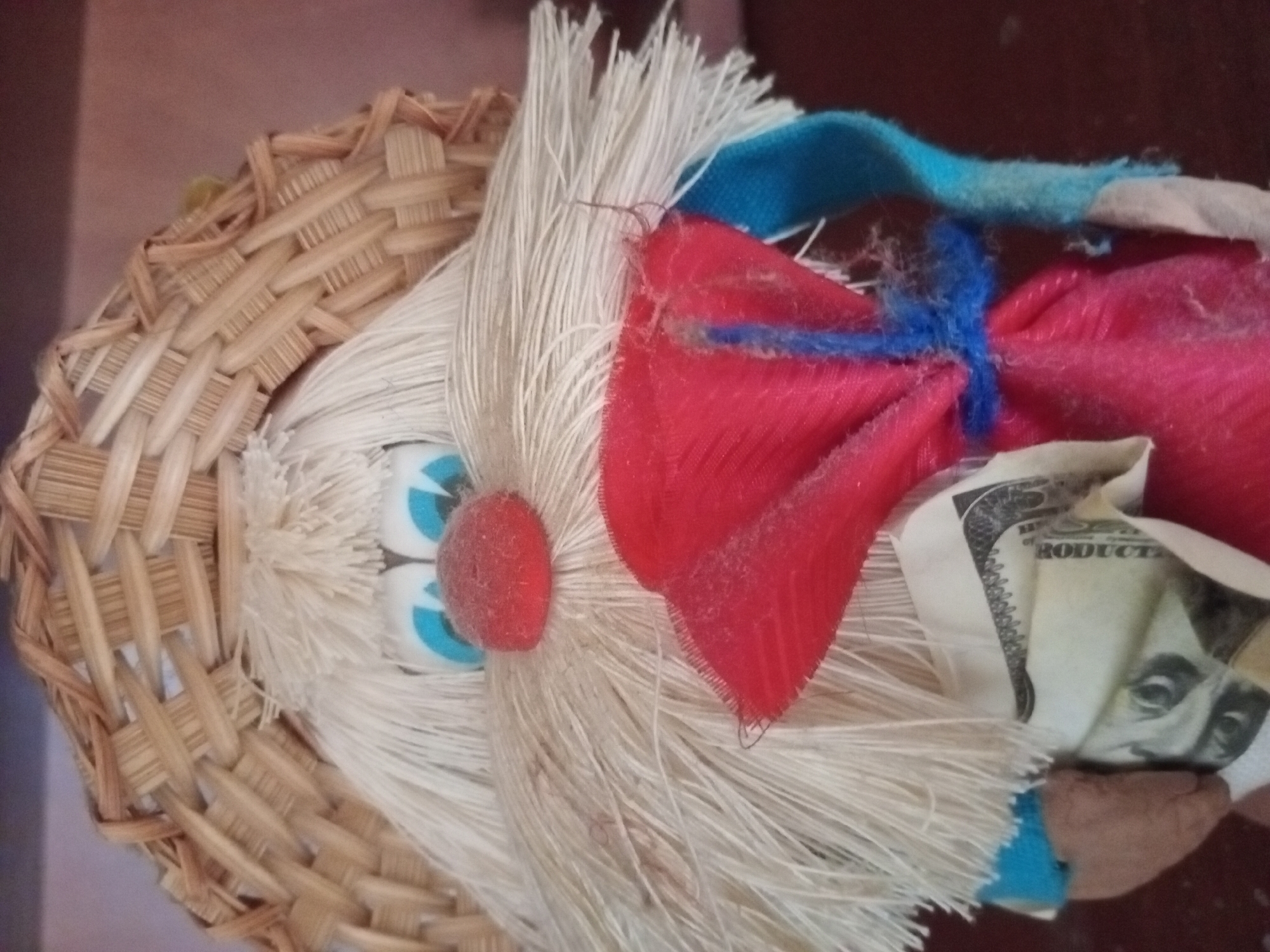 A close-up, head-on shot shows a handmade doll with a woven straw hat. The doll has a large, fluffy white beard and mustache, with large blue eyes and a red nose. It is wearing a blue shirt and a red fabric bundle tied with blue string in front of its chest. A US dollar bill is partially visible peeking out from behind the red bundle. The doll's skin-toned hands are visible on either side of the red bundle. The background is a blurred, dark brown surface.