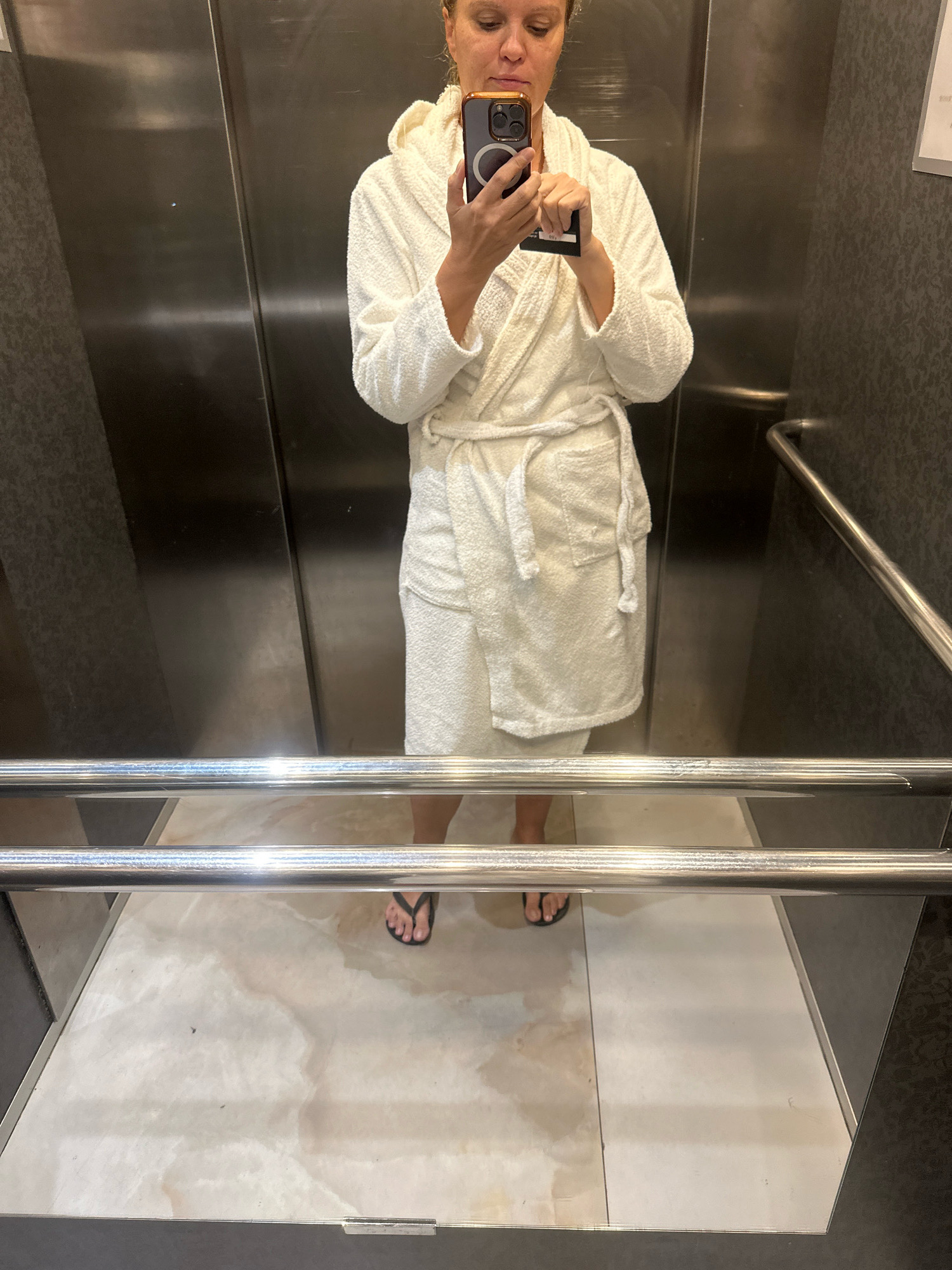 A person is standing in an elevator, taking a mirror selfie. They are wearing a white terry cloth bathrobe and black flip-flops. The elevator has stainless steel walls and a patterned gray wall. There are two silver handrails in the foreground. The floor of the elevator appears to be tiled in a light color with some darker stains. The person is holding a smartphone in their right hand, with their left hand holding a black card. The lighting is artificial and casts shadows. The location is not specified in the image or provided context.