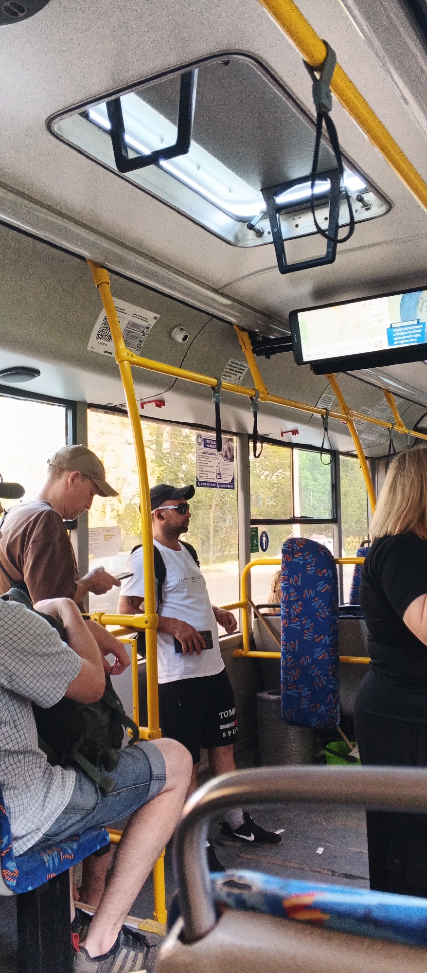 The image depicts the interior of a bus in Izhevsk, Russia. Several passengers are visible. On the left, a person wearing a cap and a brown t-shirt is looking at their phone. Next to them, a man wearing a white t-shirt with "TOMMY HILFIGER" printed on it, a black baseball cap, and sunglasses is standing and holding a phone. Further to the right, a person with blonde hair, wearing a black t-shirt, is partially visible. A blue seat with a red and blue patterned design is also visible. The bus has yellow handrails and a roof hatch with lights. A screen displaying a map is mounted above the yellow handrail. The scene appears to be during daytime, with sunlight filtering through the windows.