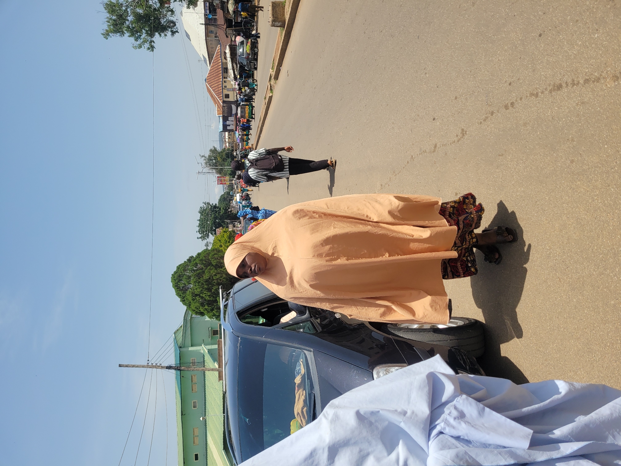 A medium shot captures a street scene in Jos, Nigeria, on a clear, sunny day. The focus is on a woman dressed in a light peach-colored hijab and patterned skirt, standing beside a dark-colored car. Her attire is voluminous, obscuring most of her body. She is looking towards her left, away from the camera. To her left, a portion of another person's light blue garment is visible, indicating their proximity. The car, a van-like vehicle, has its driver's side door ajar, and a towel or cloth is visible on the passenger seat.
The street itself is paved and appears to be an urban thoroughfare with moderate traffic. In the background, other people are walking along the sidewalk and crossing the street. A man with a backpack is walking away from the camera. Various buildings, including some with corrugated iron roofs and one prominent green building, line the street. Utility poles and wires are visible overhead, typical of an urban environment. The overall impression is that of a bustling street in a Nigerian city.