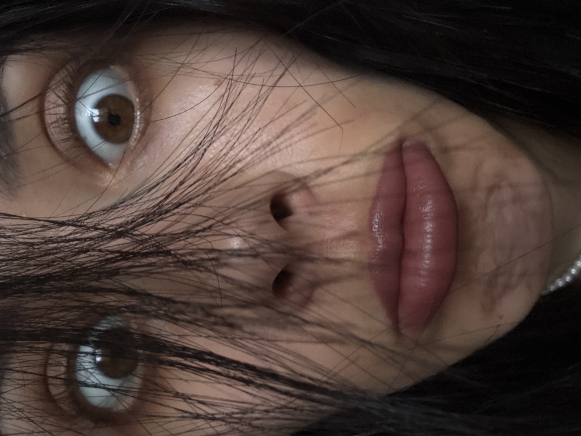 A close-up, rotated portrait shot captures the upper portion of a person's face. Dark, straight hair is spread across the face, partially obscuring the eyes and the bridge of the nose. The subject's two brown eyes are wide open, gazing directly towards the viewer with a seemingly intense or surprised expression, pupils appear dilated. The whites of the eyes are prominent. Below the eyes, part of the nose is visible, and the lips, full and a glossy reddish-pink, are slightly parted. A string of small, round, white pearl-like beads is visible along the lower right edge of the image, suggesting a necklace worn by the subject. The lighting appears soft, highlighting the texture of the lips and the individual strands of hair. No text is visible in the image. The location context for this image is Votkinsk, Russia, though no specific visual cues within the image confirm this location.
