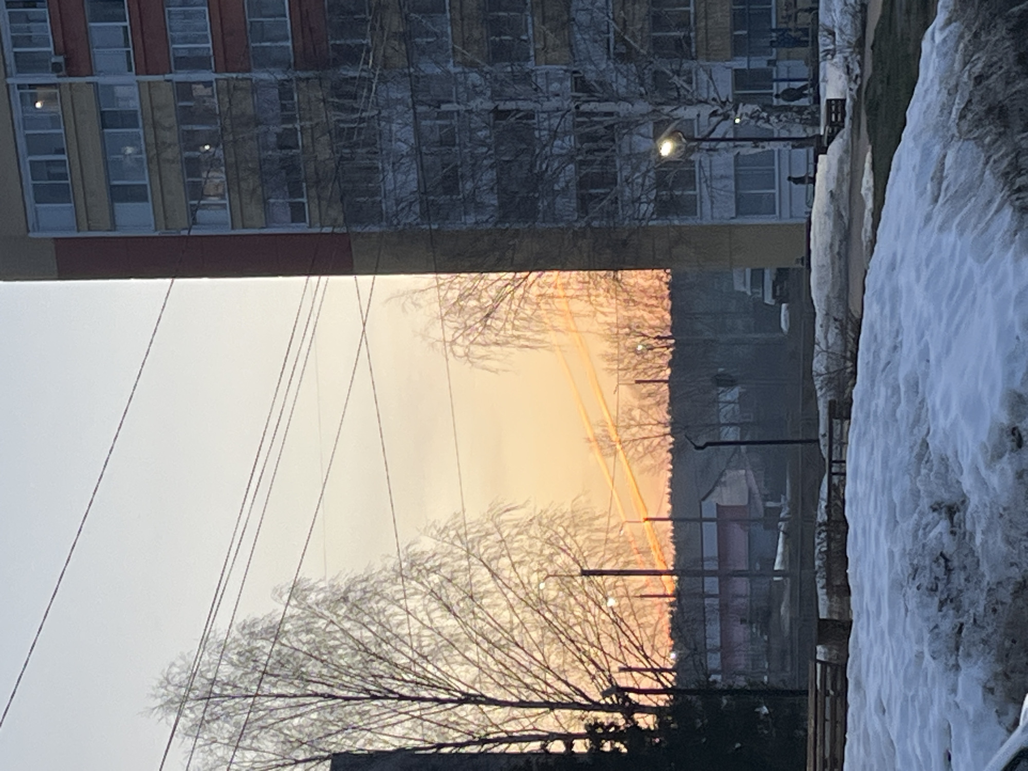 The image shows a winter scene in what appears to be a residential area of Chaikovsky, Russia. A tall building, possibly an apartment block, is on the left side of the image, with multiple rows of windows. Power lines run across the sky in the foreground. A bare tree is silhouetted against a bright, orange-colored sky, likely indicating either sunrise or sunset. To the right is a pile of snow. In the background, a building is visible. A light post can also be seen adjacent to the apartment building. The overall scene suggests a cold, urban environment.