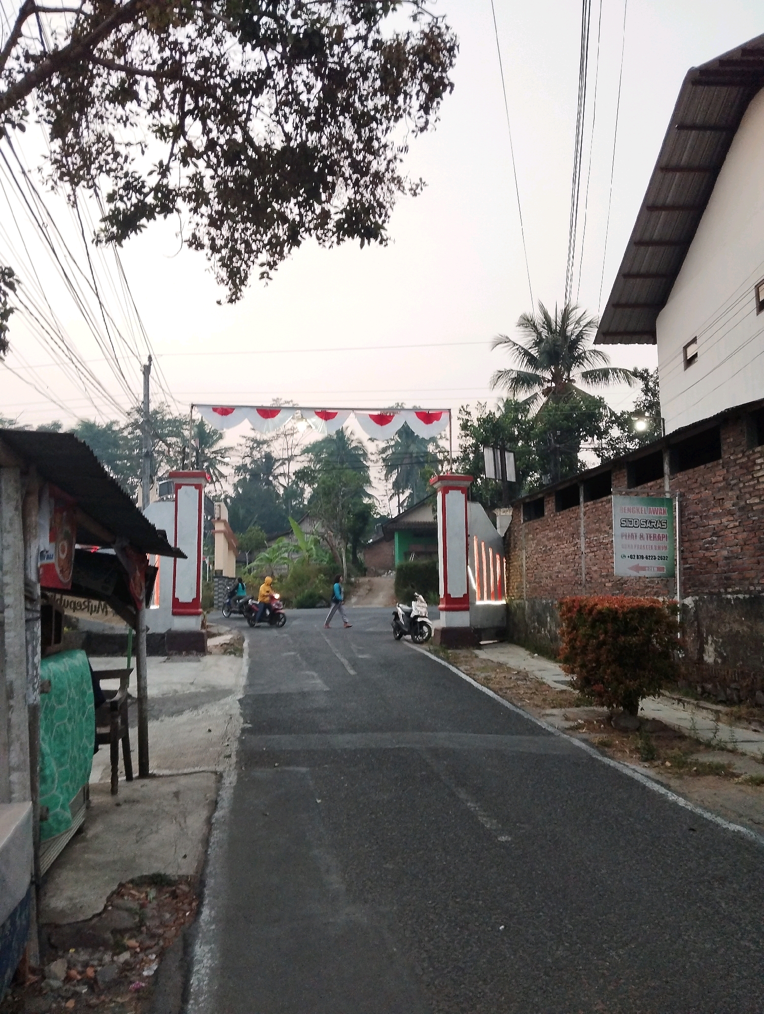 A street scene in Salatiga, Indonesia features a paved road leading towards a large white and red archway decorated with fabric banners. To the left of the road, a small, weathered structure with a corrugated metal roof and a sign is visible, partially obscuring the archway. In front of the structure, a table and chairs are set up. On the road, two motorcycles are present, one with a rider in a yellow jacket and another parked. A person in a blue shirt is walking across the road. To the right of the road, a brick wall with a white stucco top runs along the property line. A sign with Indonesian text and a phone number is attached to the brick wall. A lush green shrub with red-brown foliage is in front of the wall. The background reveals a dense collection of trees and foliage, with a palm tree standing prominently near the archway. The sky is overcast, suggesting either early morning or late afternoon. The overall atmosphere is rural or suburban.