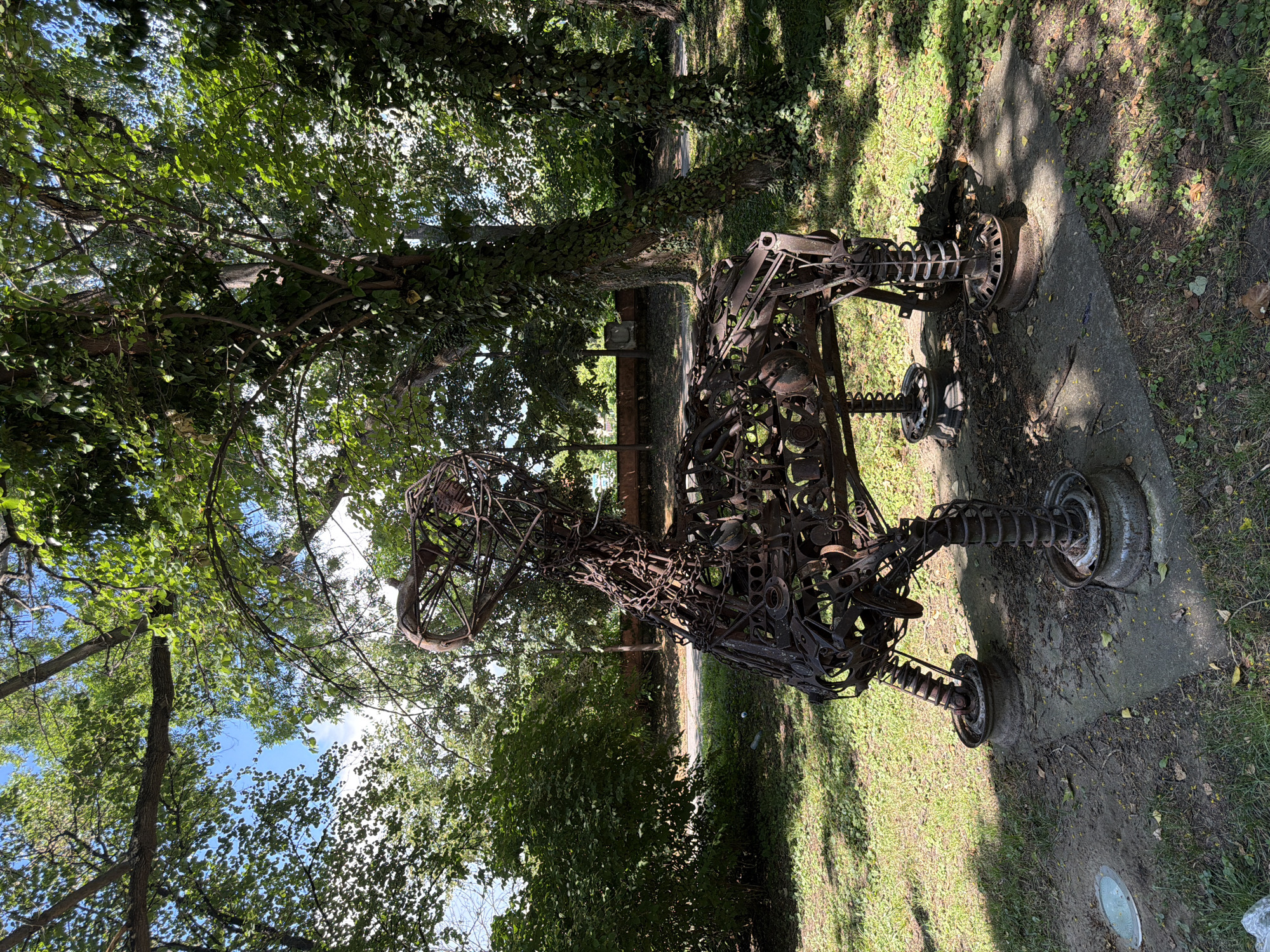 A rusted metal sculpture resembling a horse stands in a park setting. The sculpture is constructed from various metal pieces, including chains, gears, and springs, giving it a skeletal appearance. It rests on a concrete base with what appear to be repurposed metal rims as feet. The sculpture is situated in a grassy area with trees providing shade and foliage in the background. The environment suggests an outdoor public space, likely a park or garden. This artwork is located in the "Tsar Simeon" Park in Pazardzhik, Bulgaria.