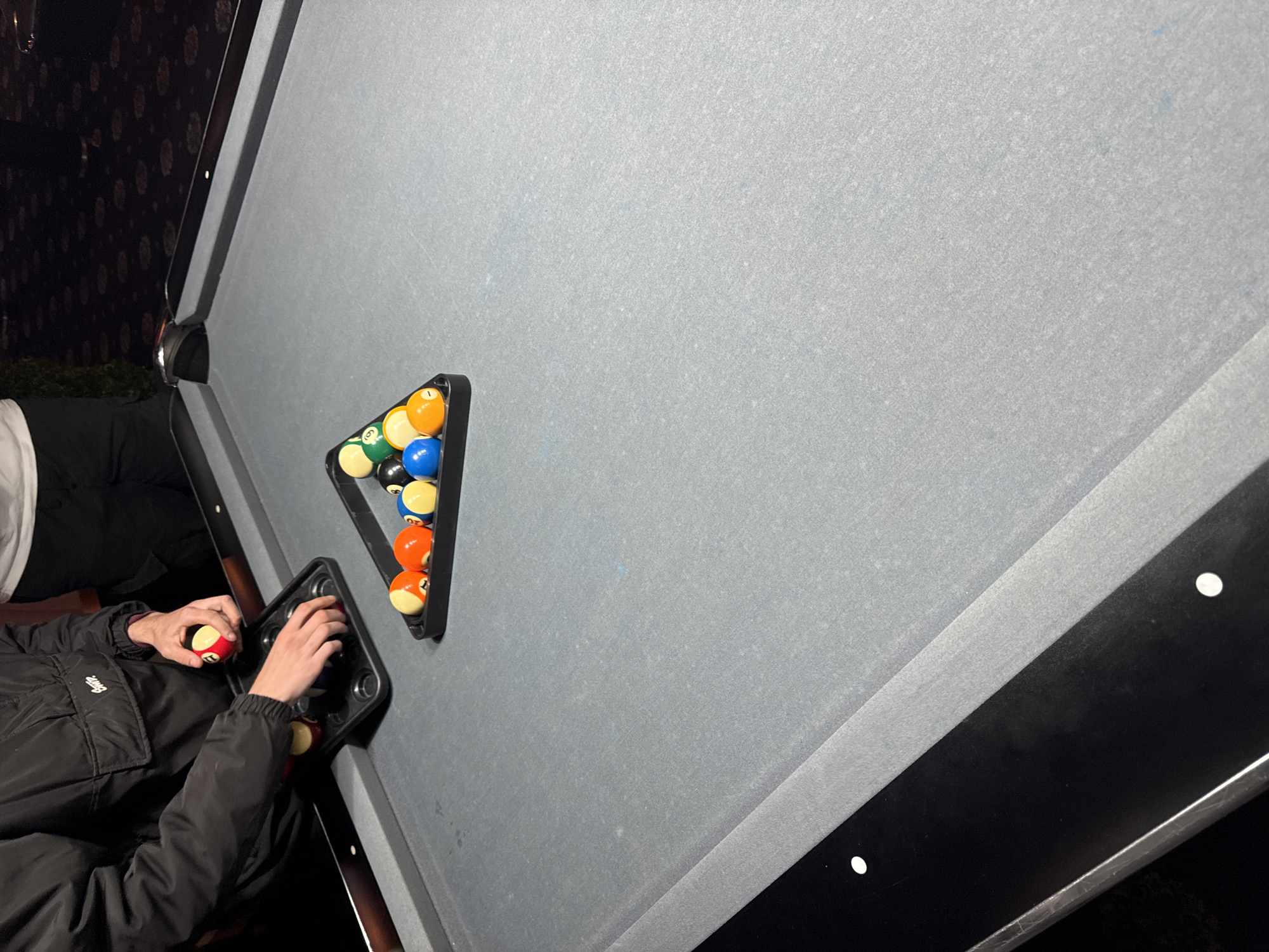 A person wearing a black jacket and a red ball is arranging the billiard balls on a billiard table. The table is grey with a black rim. The billiard balls are arranged in a triangle, ready for the game. In the background, there are plants and a wall with patterned wallpaper. The location of the image is unknown and cannot be determined.