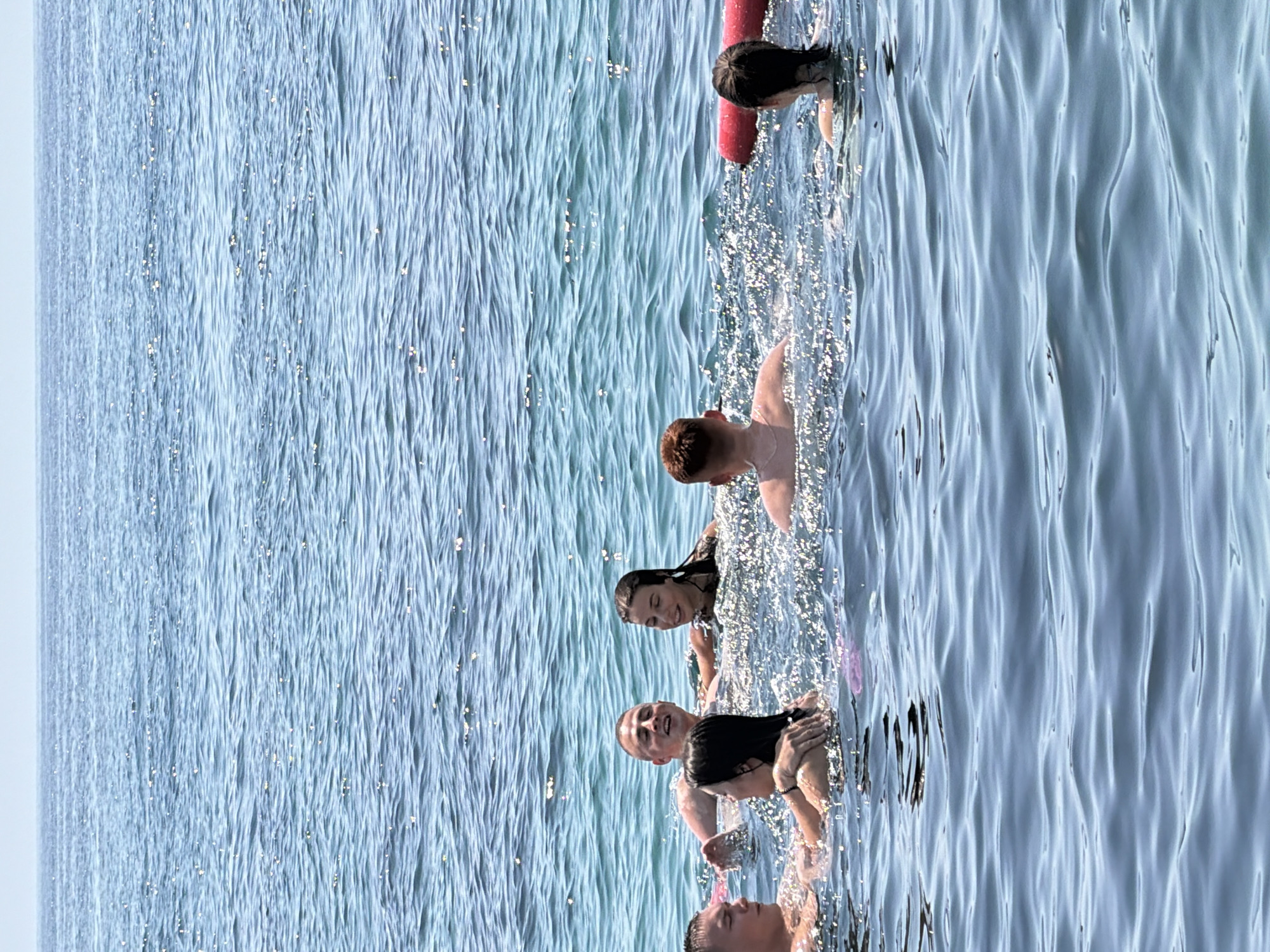 In the clear, sun-dappled waters off the coast of Budva, Montenegro, a group of seven individuals are engaged in recreational activity. Their heads and upper torsos are visible above the shimmering surface of the Adriatic Sea. The group appears to be interacting and enjoying their time together, with some individuals looking towards each other and others towards the camera. One individual on the far right is seen holding onto a red flotation device. The water is a vibrant blue, with gentle ripples and highlights from the sunlight, indicating a calm and pleasant day. The horizon is visible in the distance, a pale blue line separating the sea from the sky.