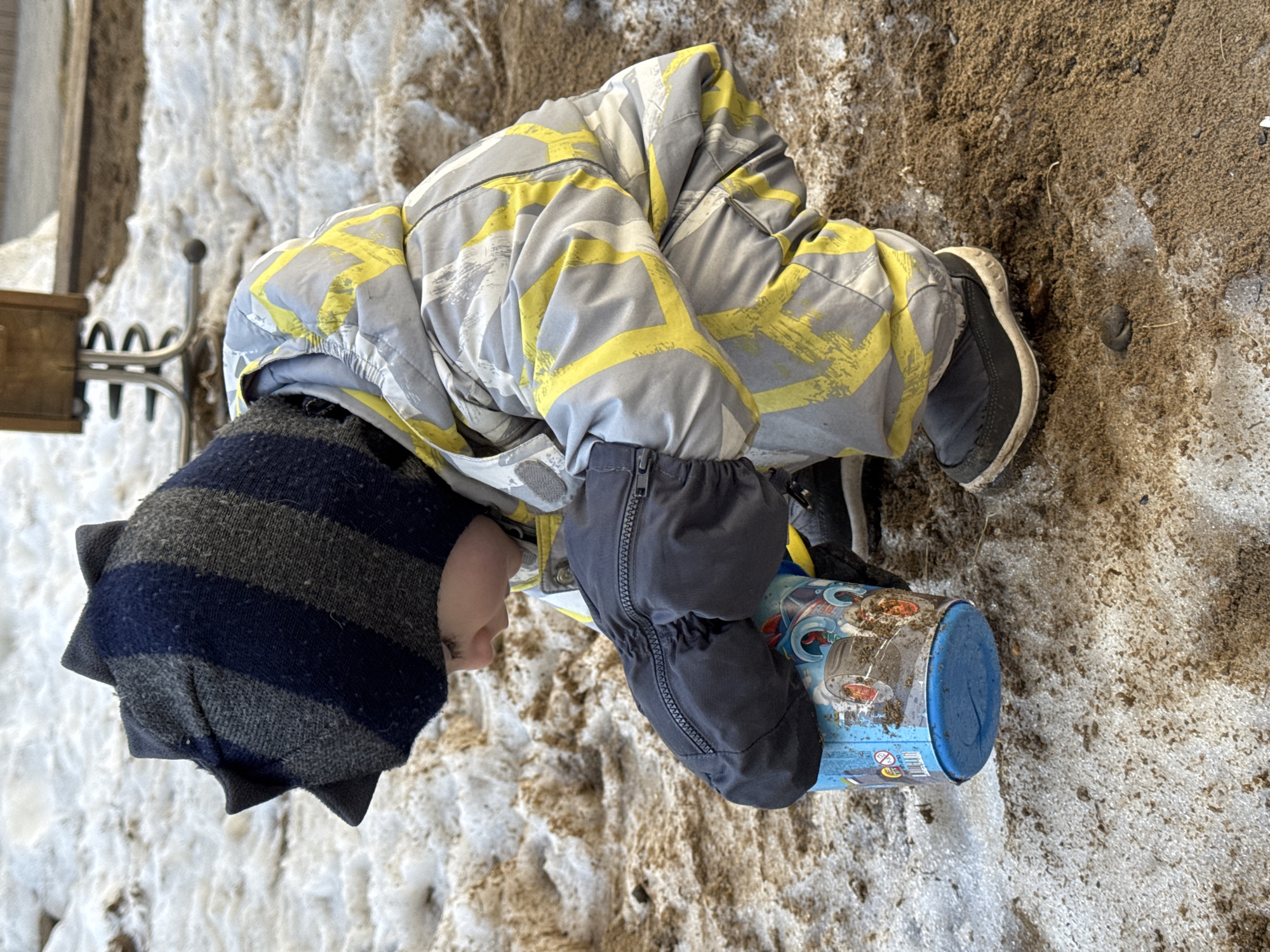 A child wearing a gray, yellow, and white patterned snowsuit, a striped blue and gray hat with spikes, and dark gray mittens is kneeling in a mixture of snow and dirt. They are holding a blue and white cylindrical container. The location appears to be outdoors, with some form of structure visible in the background on the left side. The image does not confirm the location is Izhevsk, Russia.