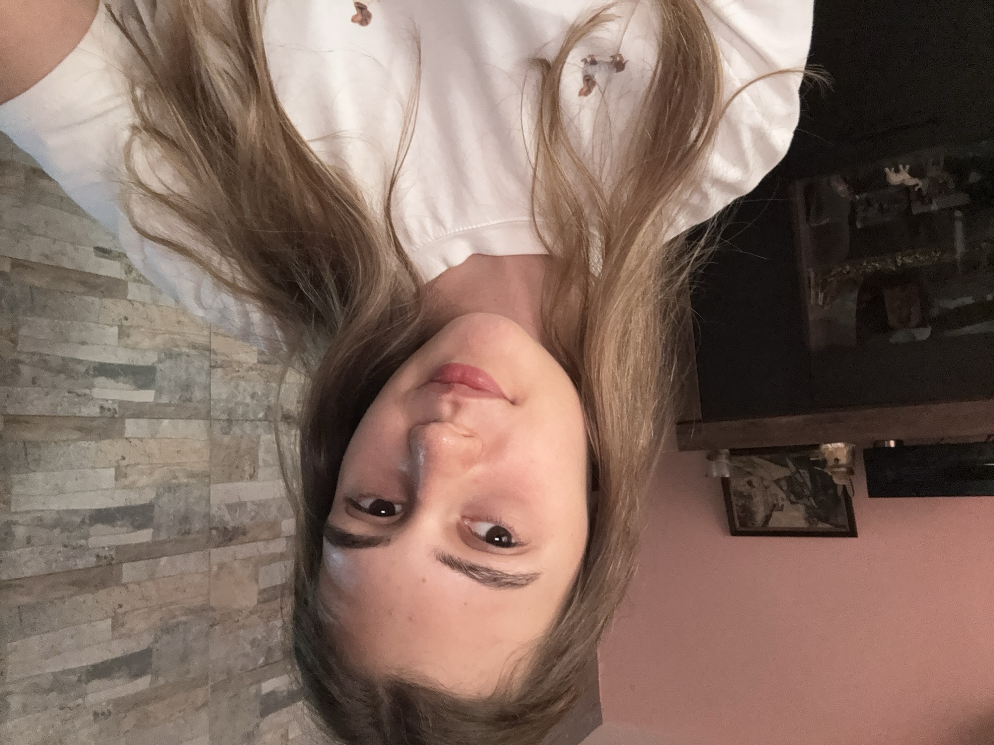 A head-and-shoulders shot captures a young woman with long, light brown hair, which appears to flow upwards due to the image being rotated 90 degrees counter-clockwise. She looks directly at the camera with a neutral, slightly contemplative expression, her lips slightly parted. She has dark eyes and well-defined eyebrows.
She is wearing a white t-shirt, visible across the upper portion of the frame (when re-oriented). The shirt features several small, irregular brownish patterns or designs.
The background on the left side of the image (when re-oriented) is a wall covered in horizontally stacked, rectangular tiles in various tones of grey, beige, and light brown. On the right, a dark, possibly wooden mantelpiece or shelf is visible. Below it, a section of a plain pink wall is seen, on which a framed picture or artwork hangs. Above the mantelpiece, some indistinct objects include a dark, rectangular screen and a small white figurine.
The scene depicts an indoor setting, likely a home, with artificial lighting. The overall impression is a casual portrait or selfie taken within a residential space in Panagyurishte, Bulgaria. There is no visible text in the image.