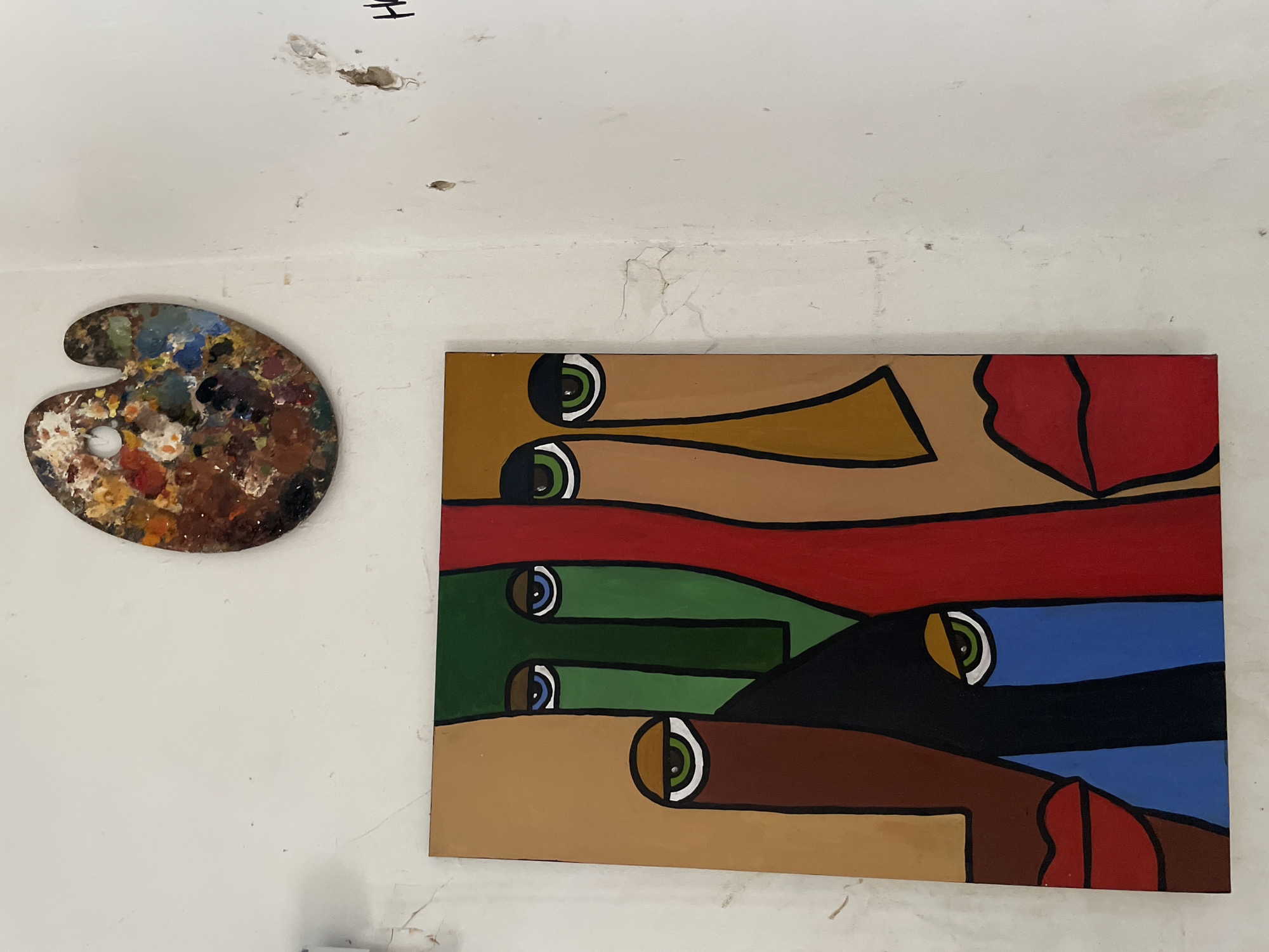 The image depicts an interior scene, likely an art studio or a home, with two distinct artistic elements on a textured, off-white wall. In the upper portion of the frame, a well-used artist's palette is mounted on the wall. It is oval-shaped and displays a rich collection of dried, multicolored oil paints, indicating active use. Below the palette, a large, abstract painting hangs on the wall. This painting features a cubist-inspired depiction of several faces, characterized by bold lines and flat planes of color. The faces are rendered in a stylized manner with prominent eyes and lips. The colors used in the painting are vibrant, including shades of tan, green, red, brown, black, and blue. The wall itself appears aged, with visible cracks and some discoloration, suggesting it might be in a developing urban environment. There is also a faint black marking on the wall to the right of the painting, possibly a tag or graffiti, that appears to spell out "H". The scene is illuminated by ambient light, creating soft shadows. Without further information or context, it is not possible to definitively locate this scene within the city of Ado, Nigeria.