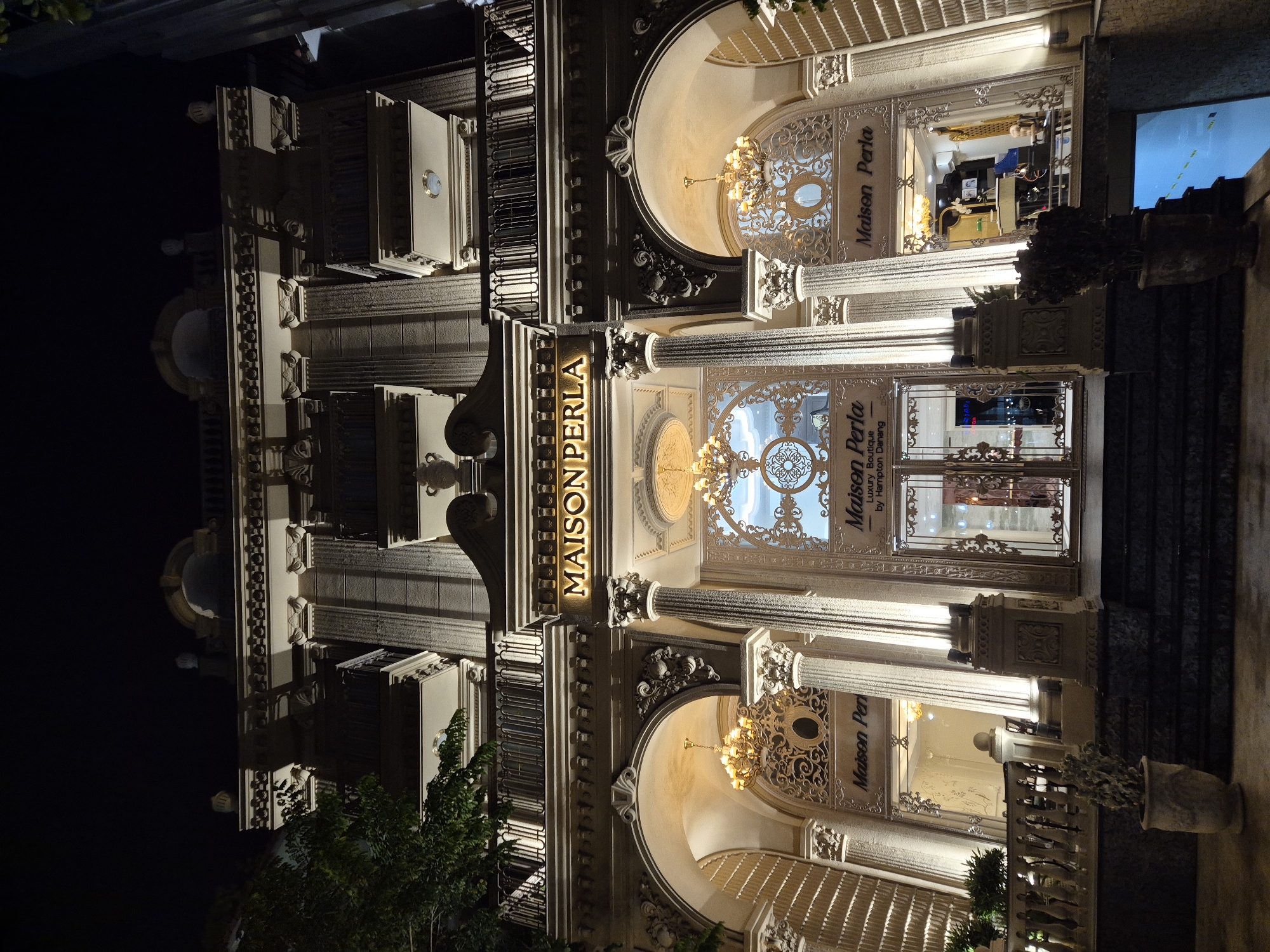 The image shows the exterior of "Maison Perla," a luxury boutique hotel in Da Nang, Vietnam, at night. The building features grand architecture with ornate columns, arched entrances, balconies, and decorative moldings, all illuminated by warm, artificial lighting. Ornate chandeliers hang from the ceilings of the arched entrances. The name "MAISON PERLA" is prominently displayed in illuminated golden letters above the central entrance. Below the name, text reads "Luxury Boutique by Hampton Danang." In the foreground, there are steps leading up to the building and potted plants. Inside the visible entrance, there are displays and furniture, suggesting a reception or lobby area. The overall scene conveys a sense of opulence and grandeur.