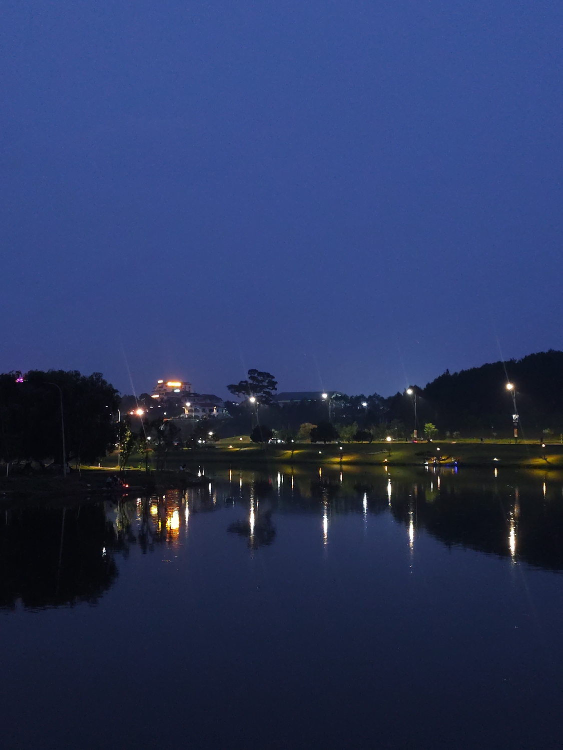 This nighttime image captures a serene view of a lake in Da Lat, Vietnam, likely Xuan Huong Lake. The scene is dominated by a dark blue sky above and a calm body of water in the foreground and midground, reflecting the numerous artificial lights from the surrounding landscape.
Along the far bank of the lake, a landscaped area with trees and a promenade is brightly illuminated by a series of streetlights. These lights create distinct, elongated reflections on the water's surface, adding a shimmering effect to the dark expanse. In the mid-right section of the lake, a small recreational boat, possibly a pedal boat, with what appears to be at least one person aboard, is visible on the water, adding a subtle element of activity.
Further in the background, on a gentle incline, a prominent building with warm yellow and orange lights stands out, suggesting a hotel or a significant establishment. The hillside is also dotted with additional trees and some distant structures. The overall atmosphere is peaceful and tranquil, characteristic of an evening stroll or relaxed evening by the water. The clear night allows for sharp reflections and a good view of the illuminated surroundings. There is no visible text in the image.