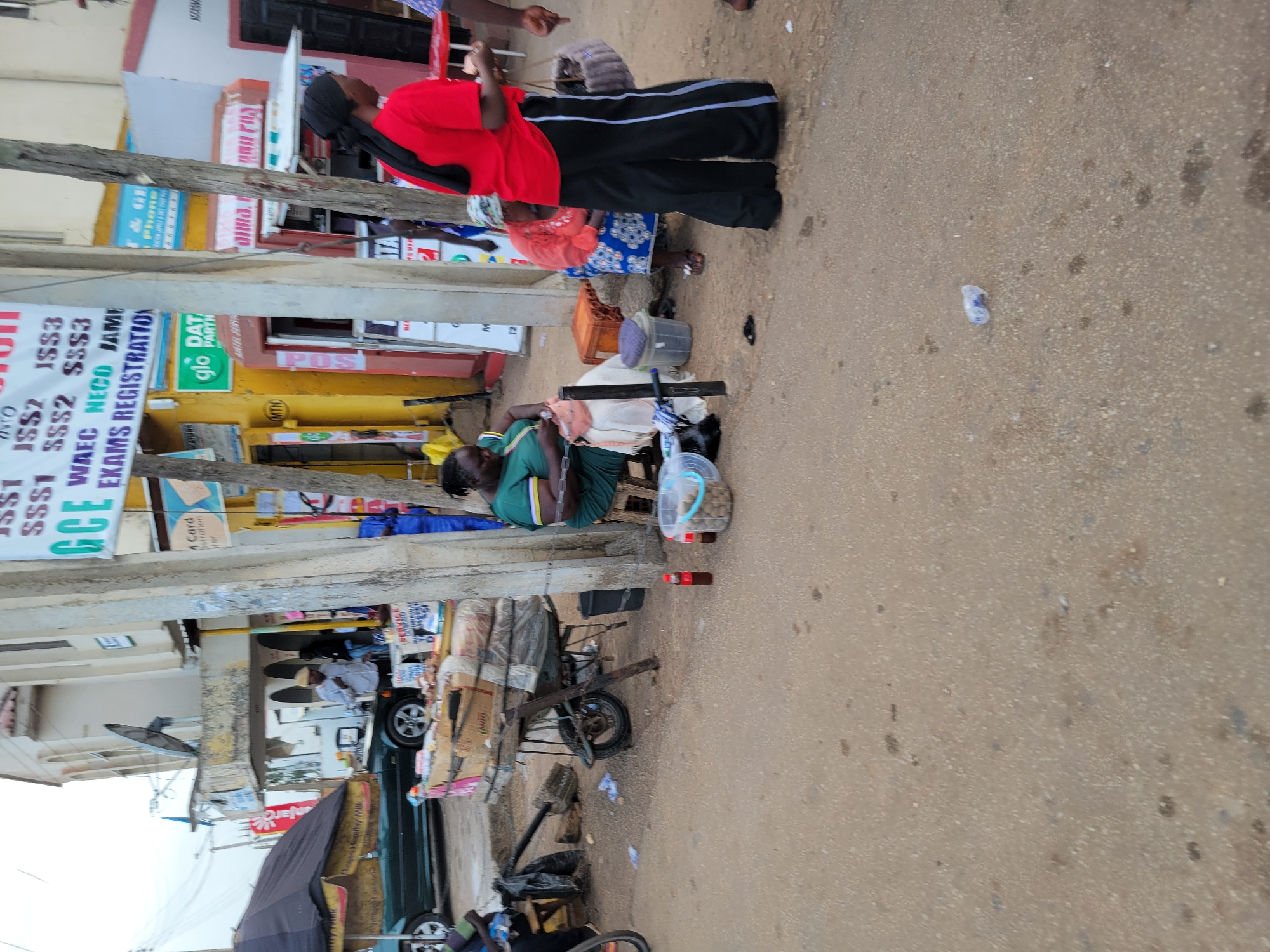 The image shows a street scene in Jos, Nigeria. In the foreground, there is a dirt road with some scattered debris. In the middle ground, a woman in a green dress is sitting on a stool, apparently engaged in some craft. Next to her is a cart filled with various goods, including what appear to be sacks of food items and boxes. Behind her, a woman in a red shirt and black pants is standing, holding a basket. Further back, there are buildings with signs, suggesting commercial activity. A dark-colored car is parked on the left. In the background, more buildings and street furniture are visible, including lampposts and what looks like a satellite dish on one of the buildings. The overall impression is of a bustling street market or commercial area.