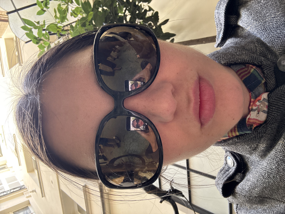 A close-up selfie captures a woman with dark hair, wearing oversized, dark-framed sunglasses, a grey textured jacket, and a multicolored plaid shirt collar. Her lips are closed. The sunglasses prominently reflect the immediate surroundings: a bicycle wheel on the left, multiple human figures walking, and a smartphone held by the subject, displaying her own reflection within the sunglasses in its screen. The background, partially visible above the woman's head, consists of light-colored, multi-story buildings and vibrant green foliage, indicating an outdoor, sunlit urban setting. The location is the city of Paris, France.