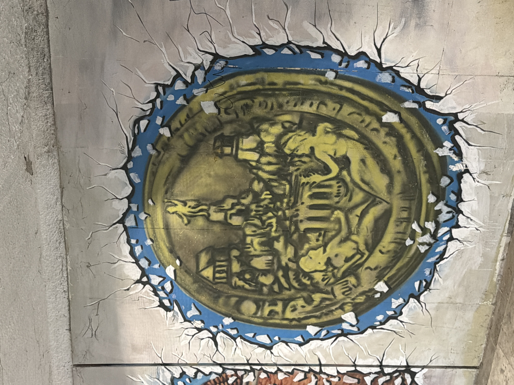 The image depicts a mural, possibly graffiti, located in Plovdiv, Bulgaria, featuring a circular design resembling a coin or medallion. The central motif is a yellow and black emblem containing figures and architectural elements that are hard to decipher. The medallion is surrounded by a blue ring, which, in turn, is bordered by a pattern of cracks that appear to be painted to create a visual effect of the medallion breaking through the surface. The background is a textured wall painted in shades of grey and white. The lower portion of the wall features a part of another mural depicting another burst with browns, whites and blacks.