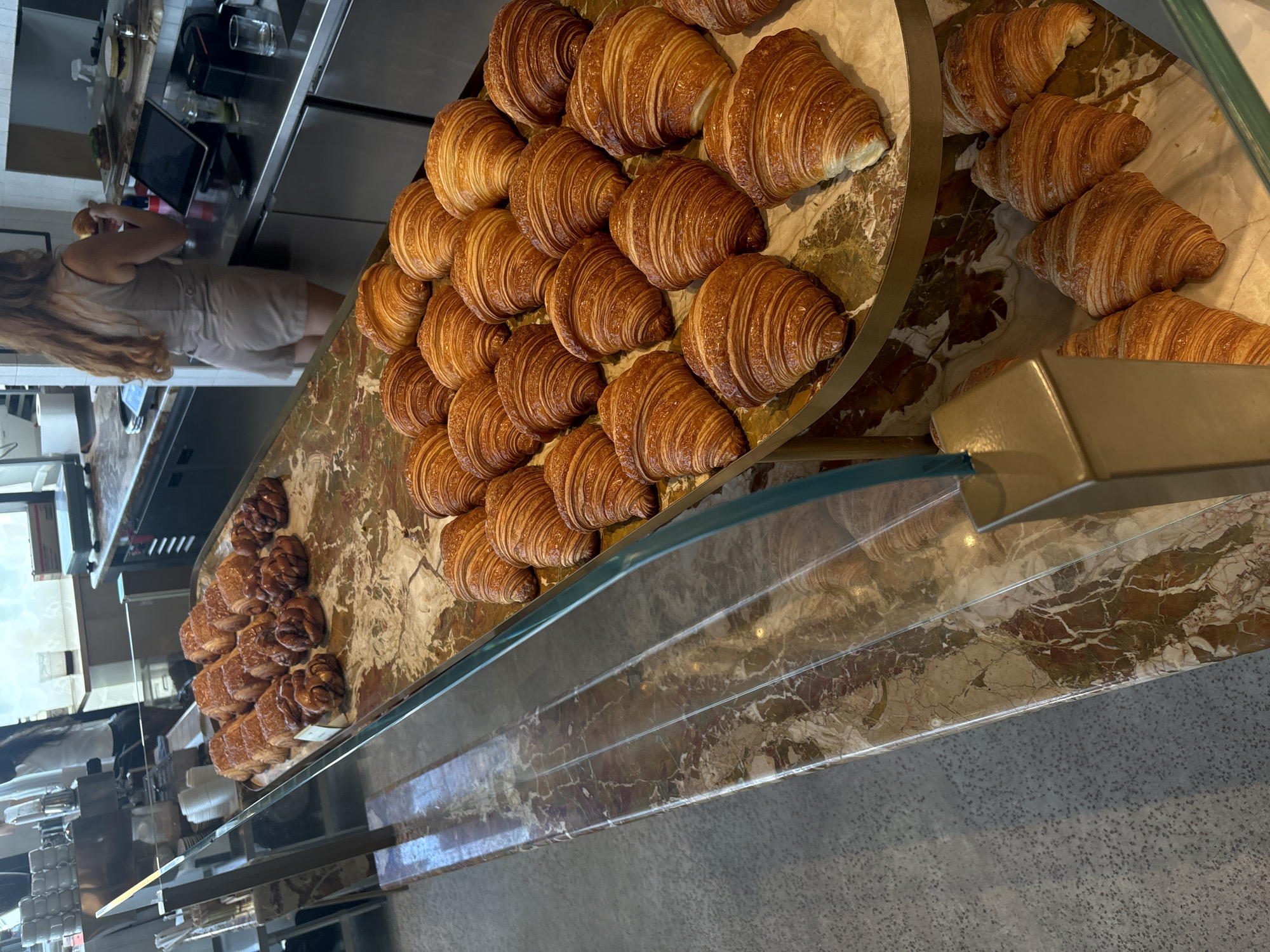A display case featuring pastries is shown at an unknown location in Miami, Florida, United States of America. The display case has a marble countertop and a glass front. Arranged on the countertop are two groupings of baked goods, specifically croissants and palmier pastries. The croissants are neatly organized on an elevated section of the countertop. To the left of the croissants, a smaller arrangement of palmier pastries is visible. A person stands behind the counter near a cash register, interacting with the display of pastries.