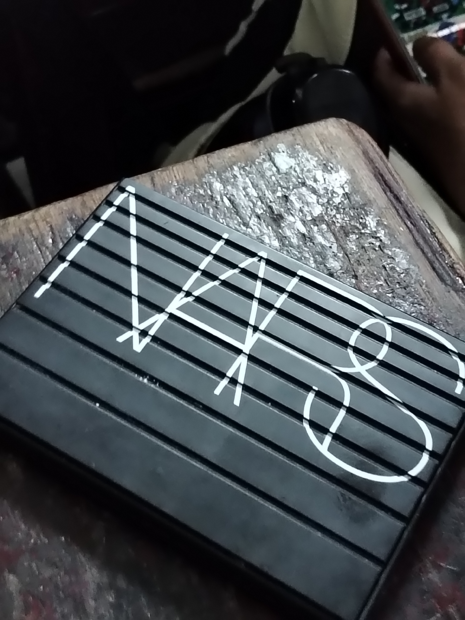 The image shows a close-up of a black makeup palette with white lettering. The palette has a ribbed surface with horizontal lines. The lettering appears to spell out "NARS," a popular makeup brand. The palette is resting on a wooden surface that looks rough and weathered, possibly a workbench or a table. In the background, out of focus, a person's hand is visible, and they appear to be holding or adjusting something. The overall lighting suggests an indoor setting, possibly during the day given the light reflected on the surface. The scene appears to be set in Davao City, Philippines.