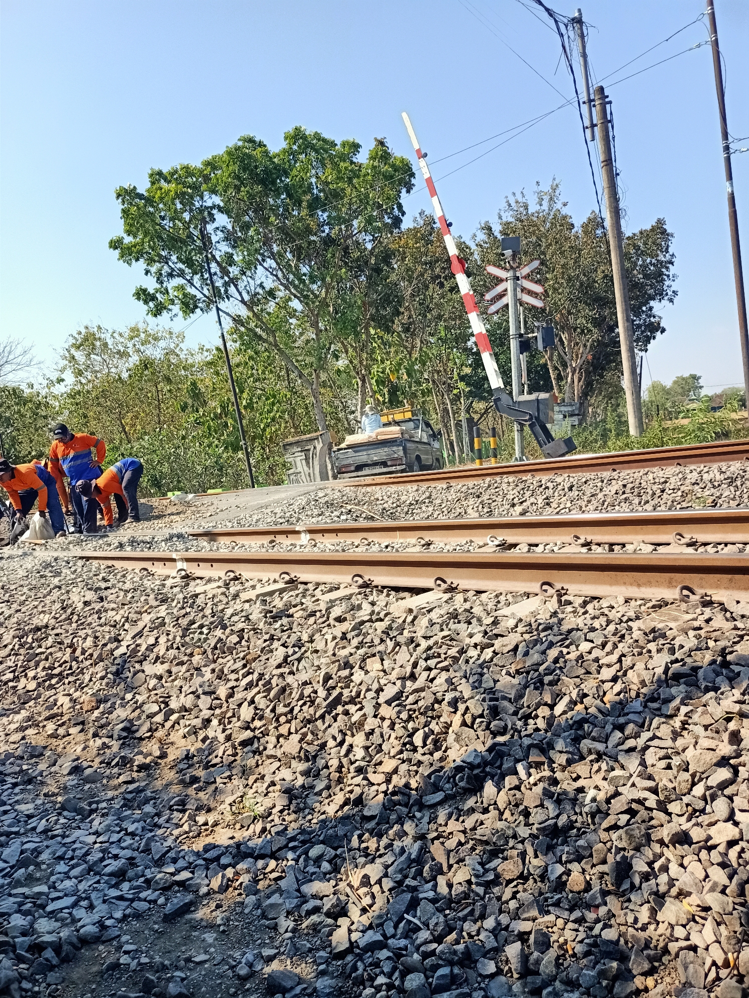 This image depicts a railway crossing in Banjarjo, Indonesia, on a clear, sunny day. Several individuals, dressed in orange work shirts and blue pants, are engaged in activities near the railway tracks. One person is bent over, appearing to be working on the ground near a white bag. Another individual in an orange shirt is standing and looking towards the tracks. A third person in an orange shirt and blue pants is also bending over near the tracks.
The railway line consists of two parallel tracks, covered with ballast stones. A railway crossing gate, painted with red and white stripes, is visible in the upright position, indicating the crossing is open. Nearby, a railway signal with red and white crossbars is mounted on a pole.
In the background, there are trees and foliage, with a bright blue sky above. A vehicle, possibly a pickup truck, is parked further down the tracks, carrying what appear to be bags of cement or building materials. Power lines and poles are also present, indicating the presence of electrical infrastructure. The overall scene suggests ongoing work or maintenance at the railway crossing.