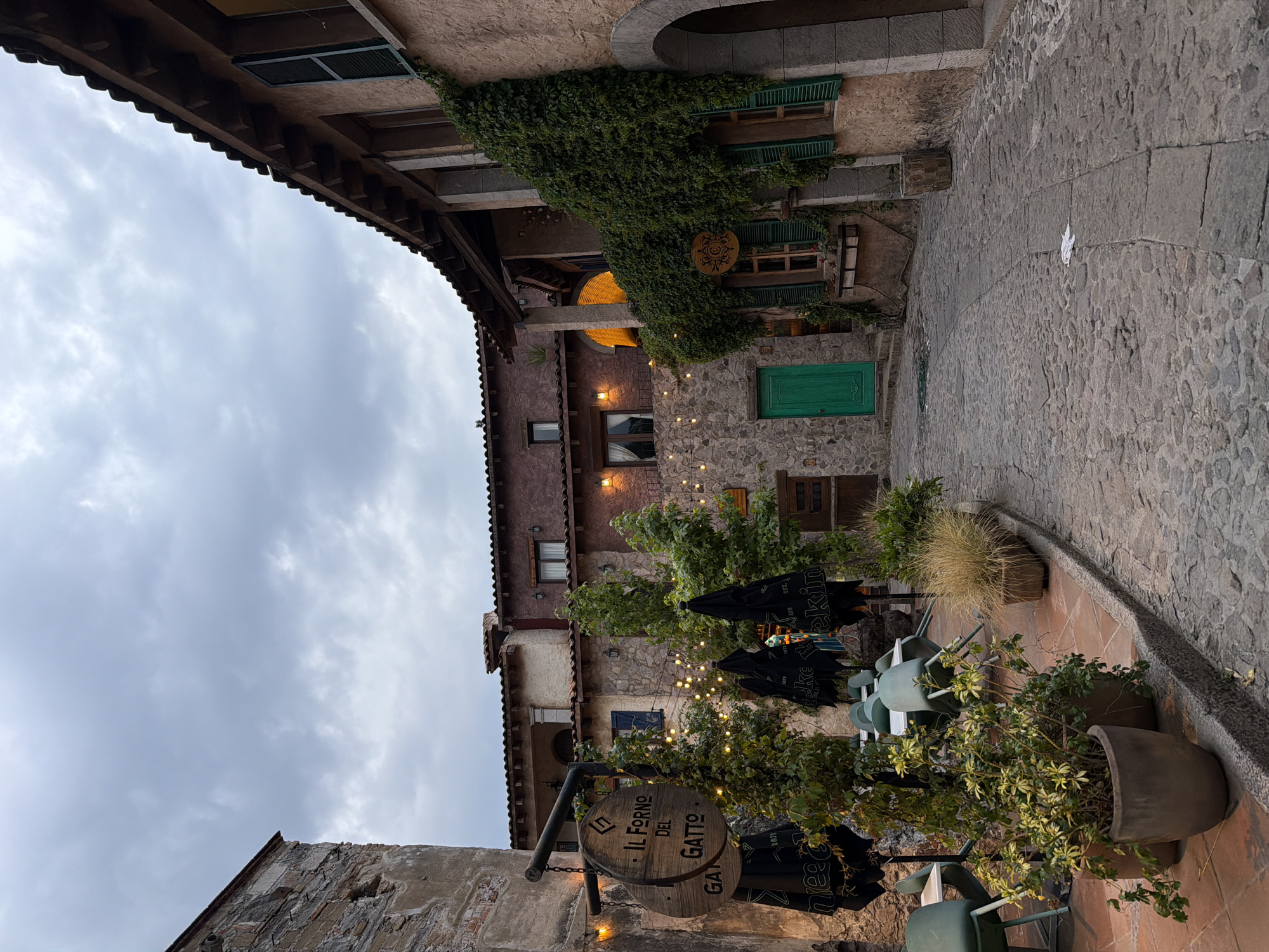 The image depicts an outdoor scene in what appears to be Domingo Arenas, Mexico. The foreground features a restaurant or cafe, suggested by tables and chairs, potted plants, and a sign that reads "IL FORNO DEL GATTO." Overhead lighting is strung between trees, creating a warm ambiance. The architecture consists of buildings made of stone, and the green door adds a rustic touch. The sky is cloudy, indicating an overcast day. The image seems to capture a casual scene in a cozy setting.