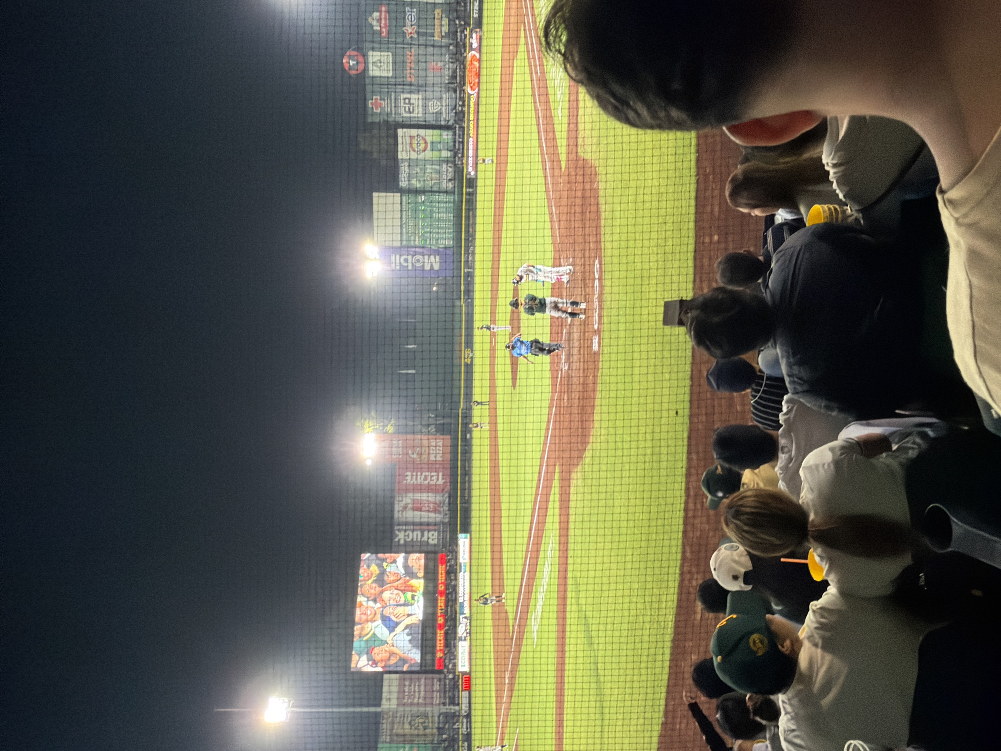 The image depicts a baseball game at night. Spectators are in the foreground. The field is visible through a net. The batter's box has a batter, a catcher, and an umpire. A second batter is on the on-deck circle. A third batter is standing on the first base. Various advertisements, including Mobil and Tecate, are visible on the stadium walls. A large video screen is showing images of people. The location is likely in the city of Puebla, Mexico, given the language on the billboards and the uniform details of the players.
