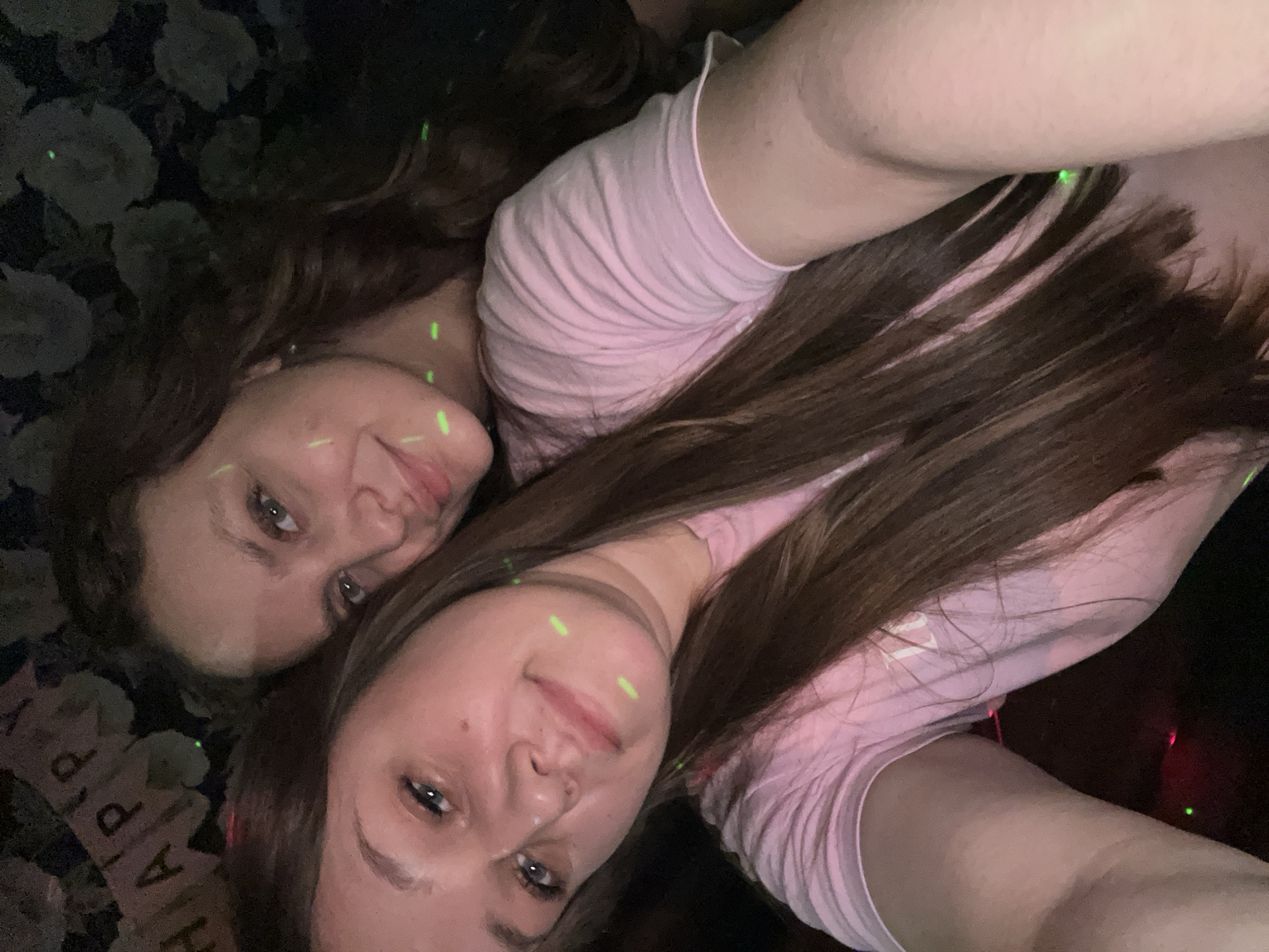 Two young adult females are depicted in a close-up indoor selfie, located in the city of Ольховка, Russia. The female in the foreground, with long brown hair and a pink t-shirt, holds the camera, looking directly at the viewer with a slight smile. Her face and arm show several distinct luminous green light specks. Behind her, a second female leans into the frame, also smiling towards the camera. She has long dark brown wavy hair and green or blue eyes, with similar green light specks visible on her face. The background is dimly lit, revealing a dark wall with a subtle floral pattern. In the upper left quadrant, a portion of a "HAPPY" banner is visible, suggesting a celebratory event. The presence of laser-like green light specks on the individuals and in the environment indicates a party or club setting.