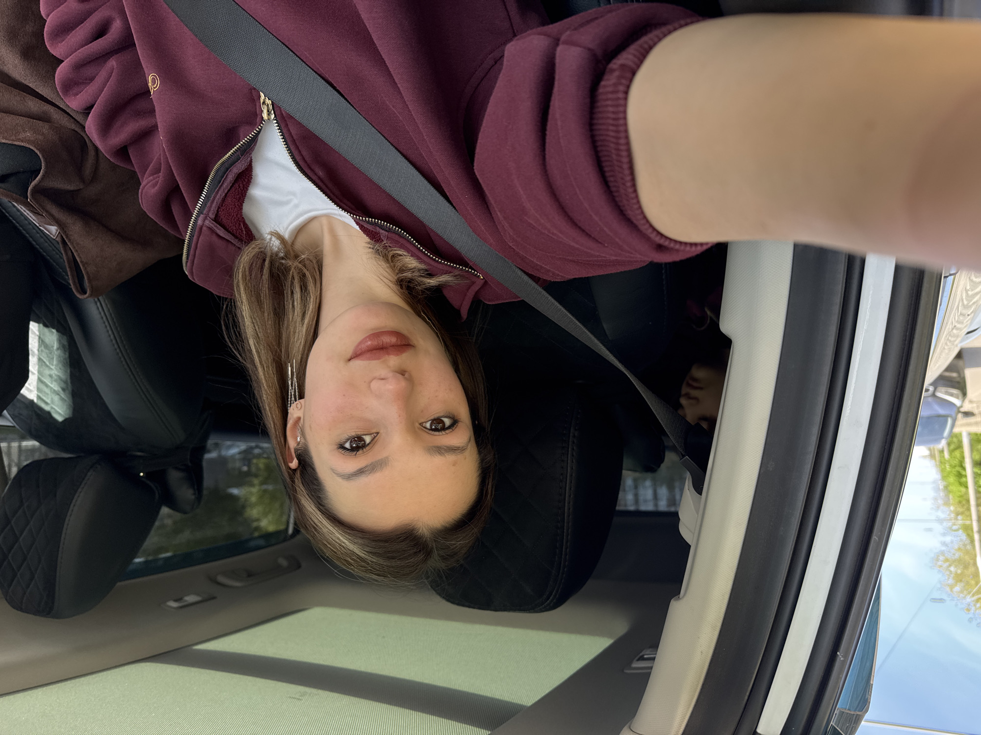 The image is a selfie taken inside a car, rotated 90 degrees counter-clockwise. The primary subject is a young woman, seen from the chest up, looking directly at the camera. She has medium-length brown hair with lighter highlights, brown eyes, and is wearing light makeup including lipstick and filled-in eyebrows. On her visible left ear, she wears two silver earrings: a small hoop and a longer, dangling chain-like earring.
She is wearing a maroon zip-up hoodie over a white t-shirt, with the zipper partially open. A black seatbelt diagonally crosses her chest. On the upper right chest area of her hoodie, a faint gold-colored, unidentifiable logo or text is visible.
The setting is the interior of a car. Black leather seats are visible behind her and to her left, with one seat headrest featuring a quilted pattern. Through the car windows and sunroof, natural daylight is visible, suggesting it is daytime and likely sunny, with some trees visible outside. In the background, partially obscured by shadow and a car seat, the head of another person, possibly a child, can be seen in the back seat. The car's interior trim, including light-colored plastic panels and dark rubber seals around the door frame, is also visible on the right side of the frame.
The activity taking place is a selfie being taken by the woman while she is seated in the car, likely as a passenger. Her expression is calm and neutral, with a slight hint of a smile.