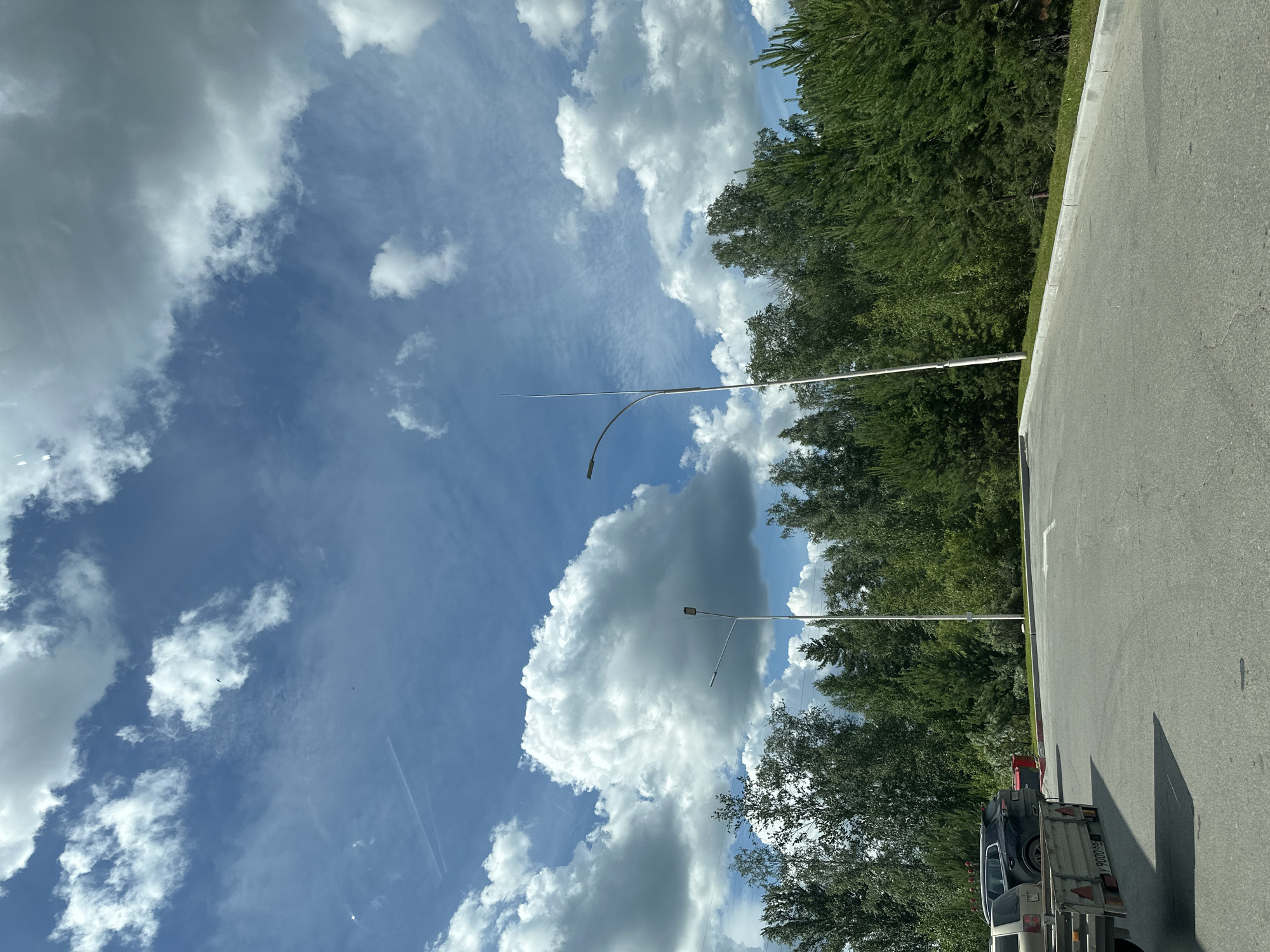 The image shows a street with trees and a blue sky with clouds. There are cars parked on the side of the road, and a trailer is visible in the foreground. Two lampposts are present, one in the foreground and one in the background. The scene appears to be a quiet, possibly suburban or rural, area. The weather is sunny with scattered clouds. There is a white arrow painted on the asphalt, indicating a turn. The image was captured in Yekaterinburg, Russia.